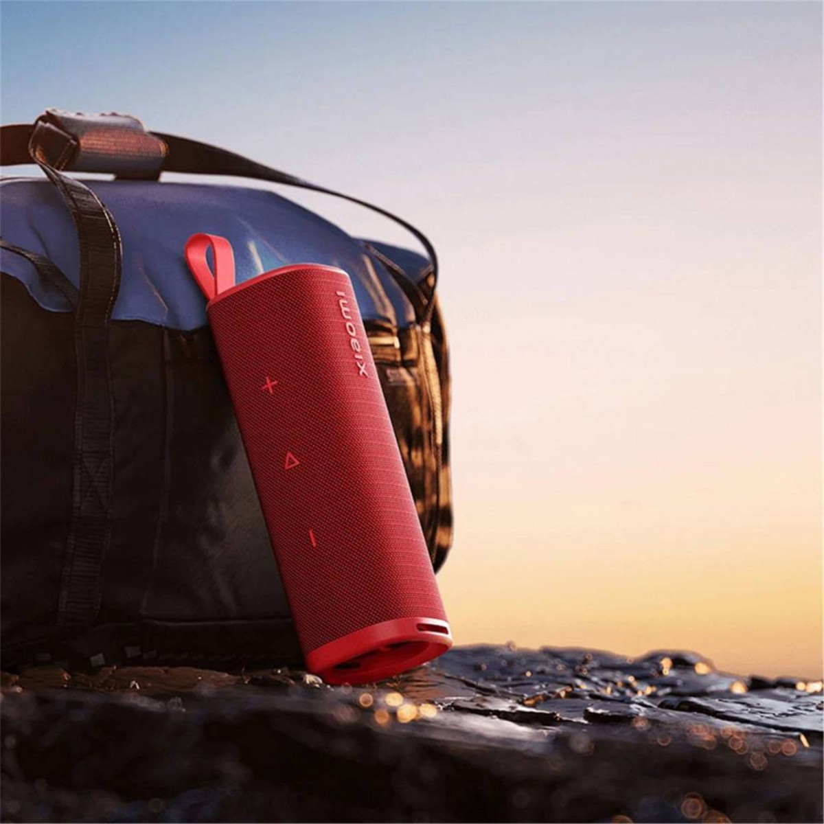 Czerwony głośnik Xiaomi obok niebiesko-czarnej torby, na tle zachodu słońca z wodą i niebem.
