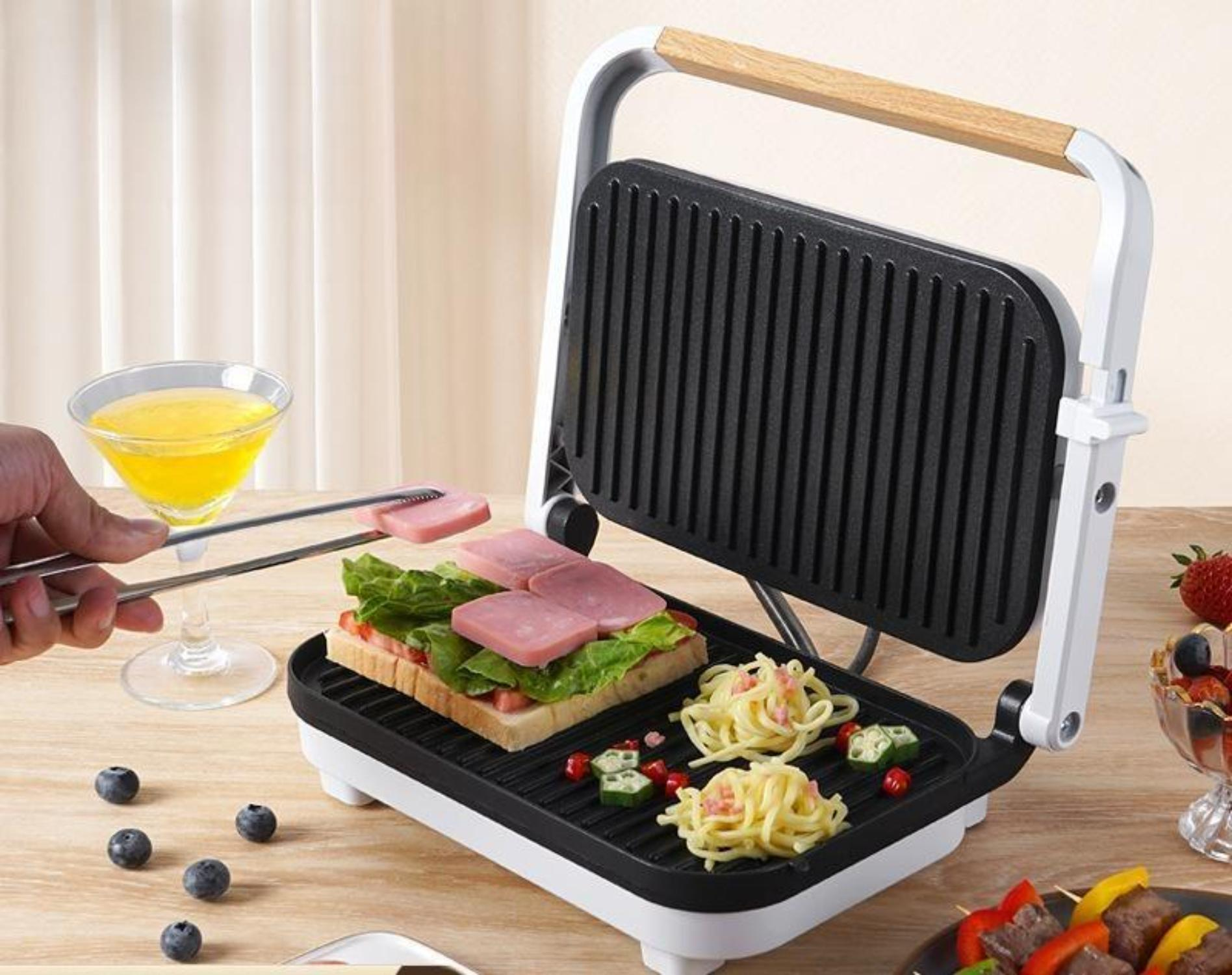 Panini-Presse mit Essen darin und eine Person, die mit Zangen Essen hinzufügt.