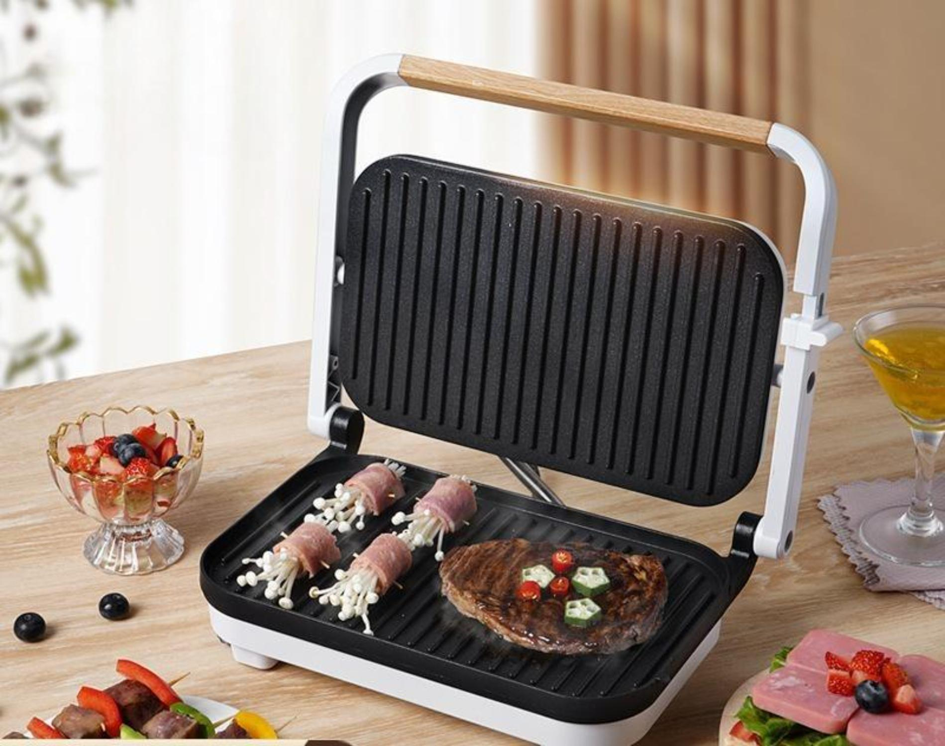 Panini-Presse mit Essen, einschließlich Steak, umgeben von Beilagen. Heller Hintergrund.
