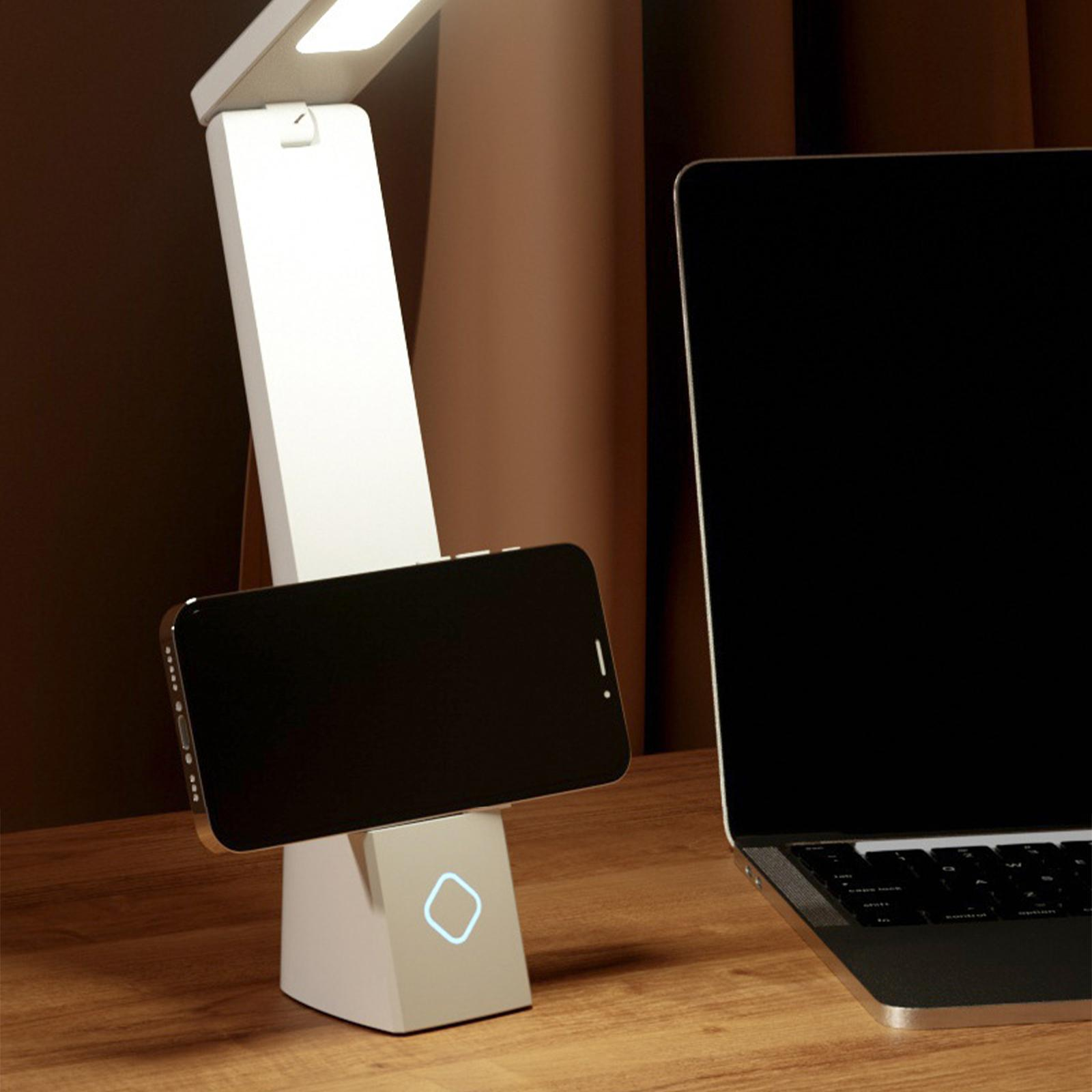 Eine weiße Schreibtischlampe mit Telefon auf dem Fuß. Ein Laptop im Hintergrund, auf brauner Oberfläche.