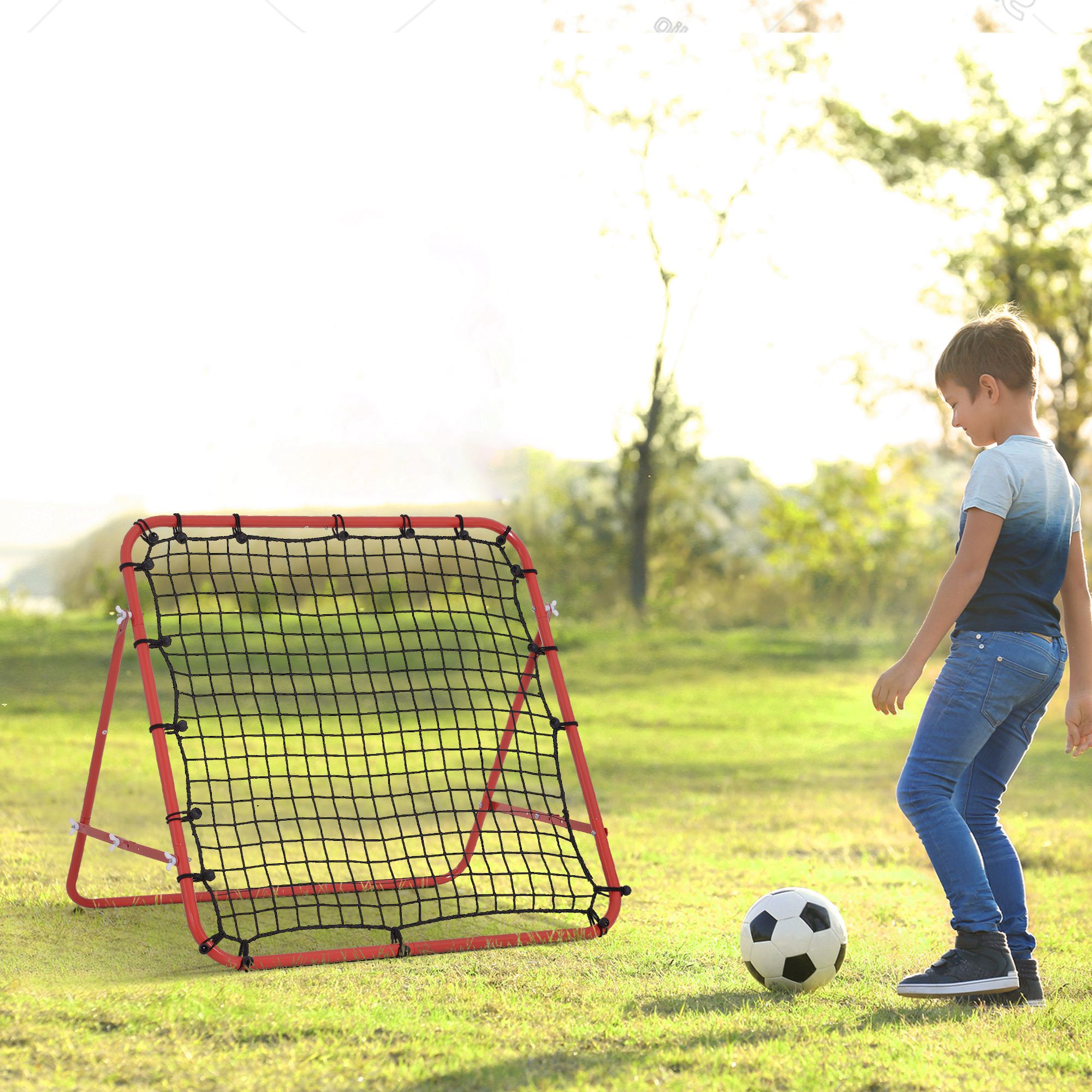 Ein Junge tritt einen Fußball in Richtung eines roten Rebounders auf einem grünen Rasen unter hellem Himmel.