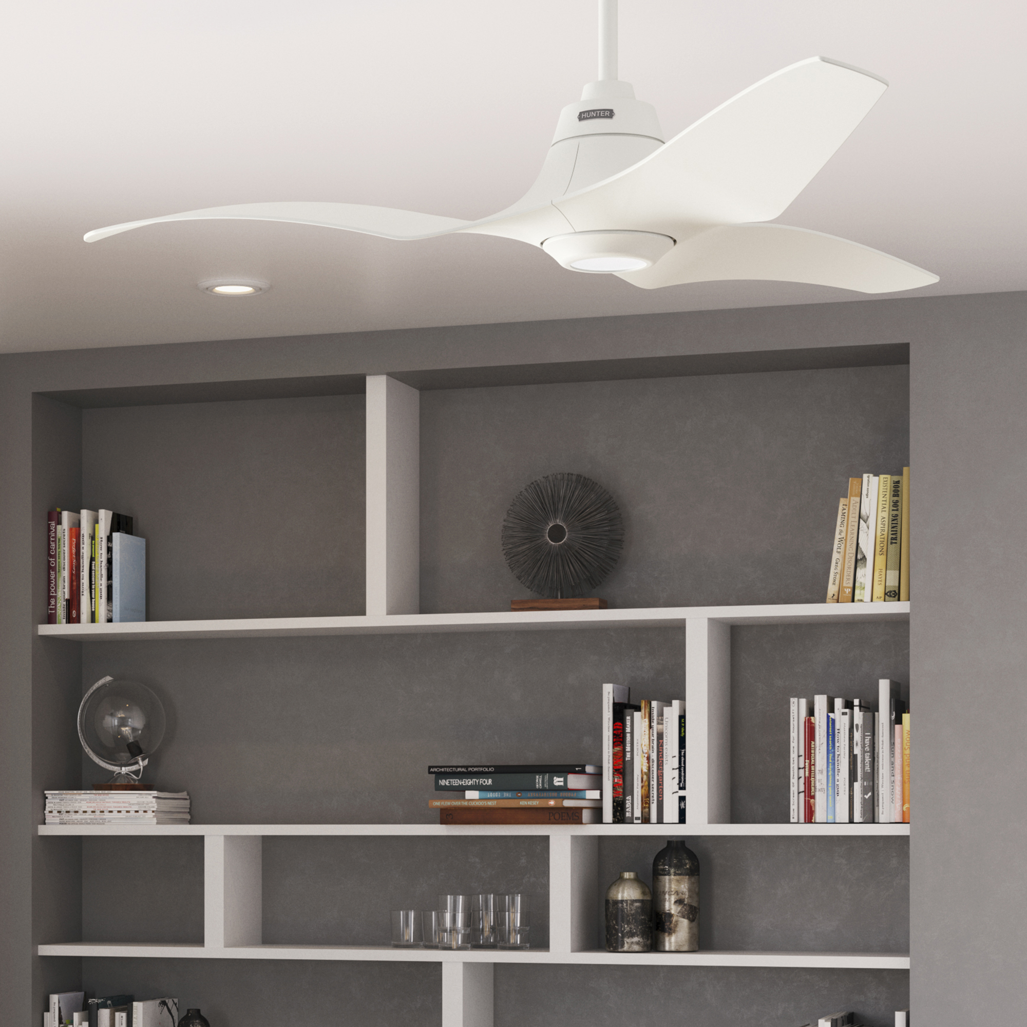Ein weißer Deckenventilator mit drei Blättern. Der Hintergrund zeigt eingebaute Regale und eine dunkelgraue Wand.