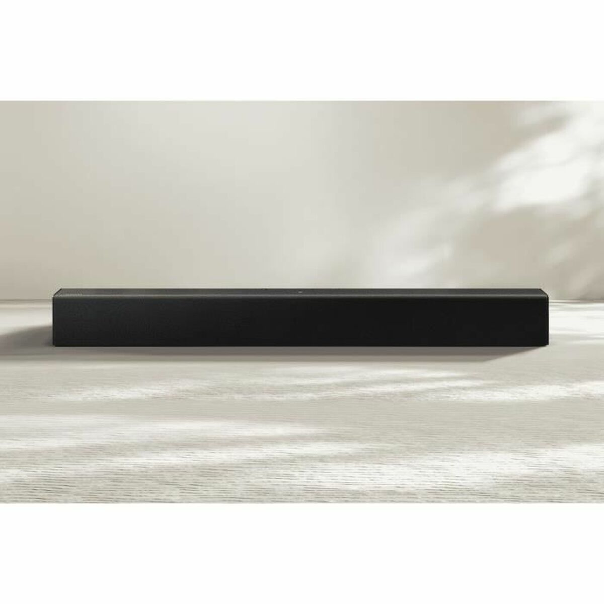 Eine schwarze Soundbar auf einer hellen Holzoberfläche. Der Hintergrund ist eine helle Wand mit Schatten.