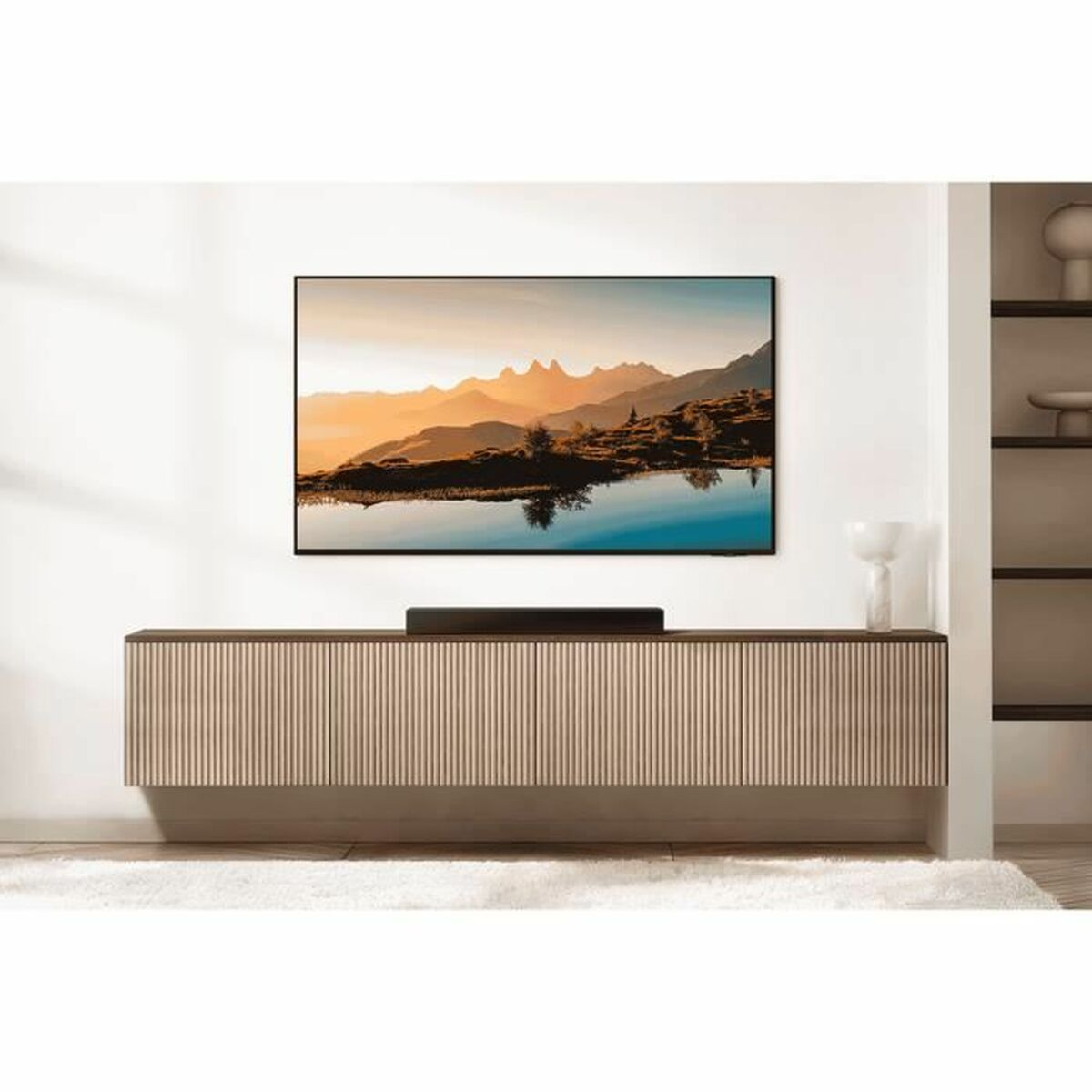 Ein Fernseher zeigt Berge über einem See. Der Fernseher steht auf einem Holzschrank mit einer Soundbar. Weißer Teppich darunter.