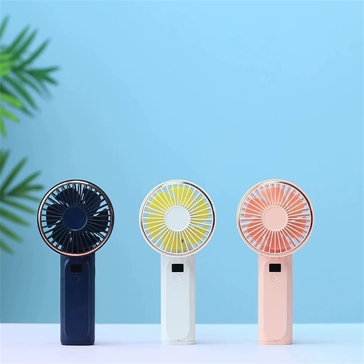 Drei verschiedenfarbige tragbare Ventilatoren in einer Reihe. Sie sind weiß, marineblau und rosa, mit goldenen Zierleisten.