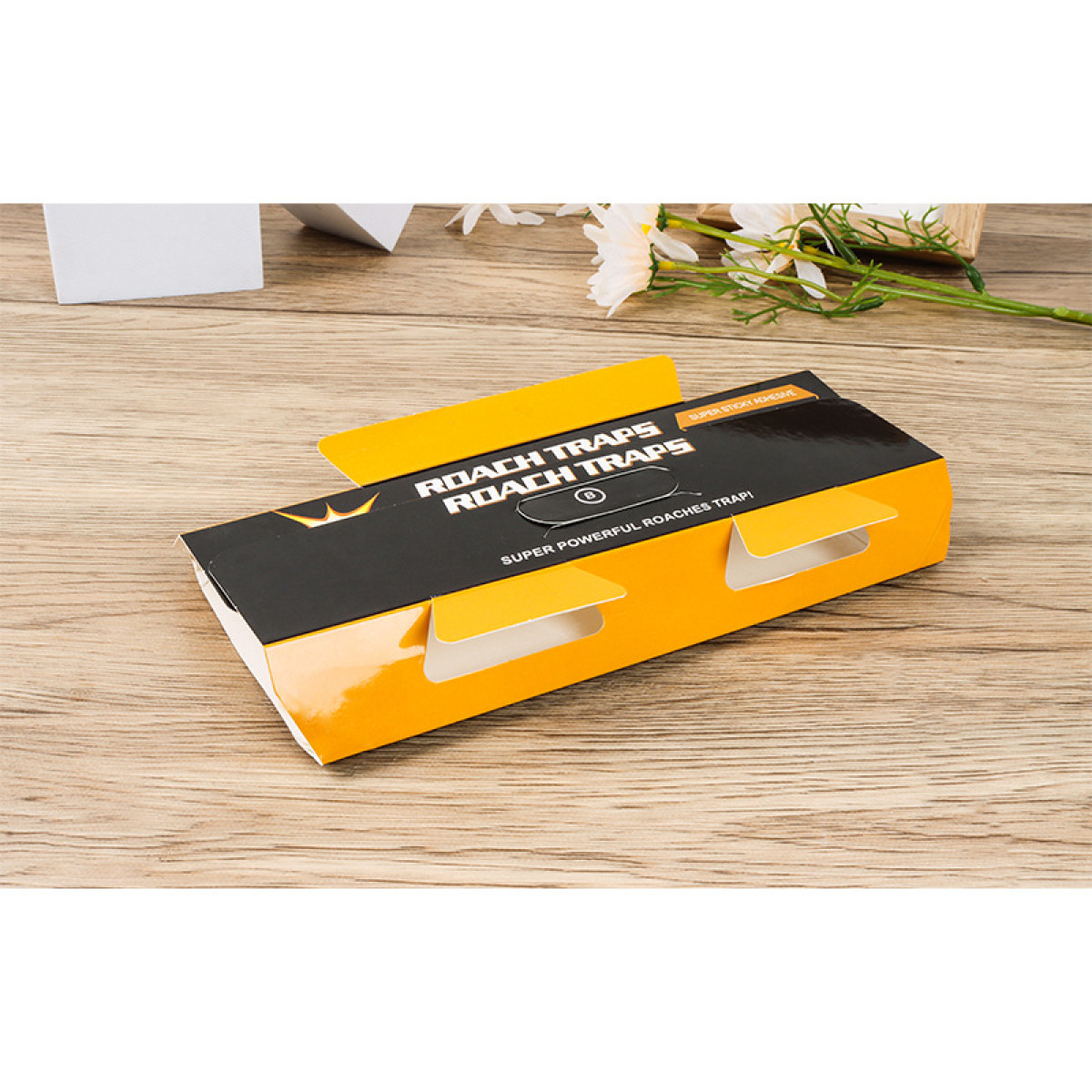 Kakerlakenfallen-Verpackung offen auf einem Holztisch. Die Falle ist schwarz und gold, mit Text auf der Verpackung. Weiße Blumen.