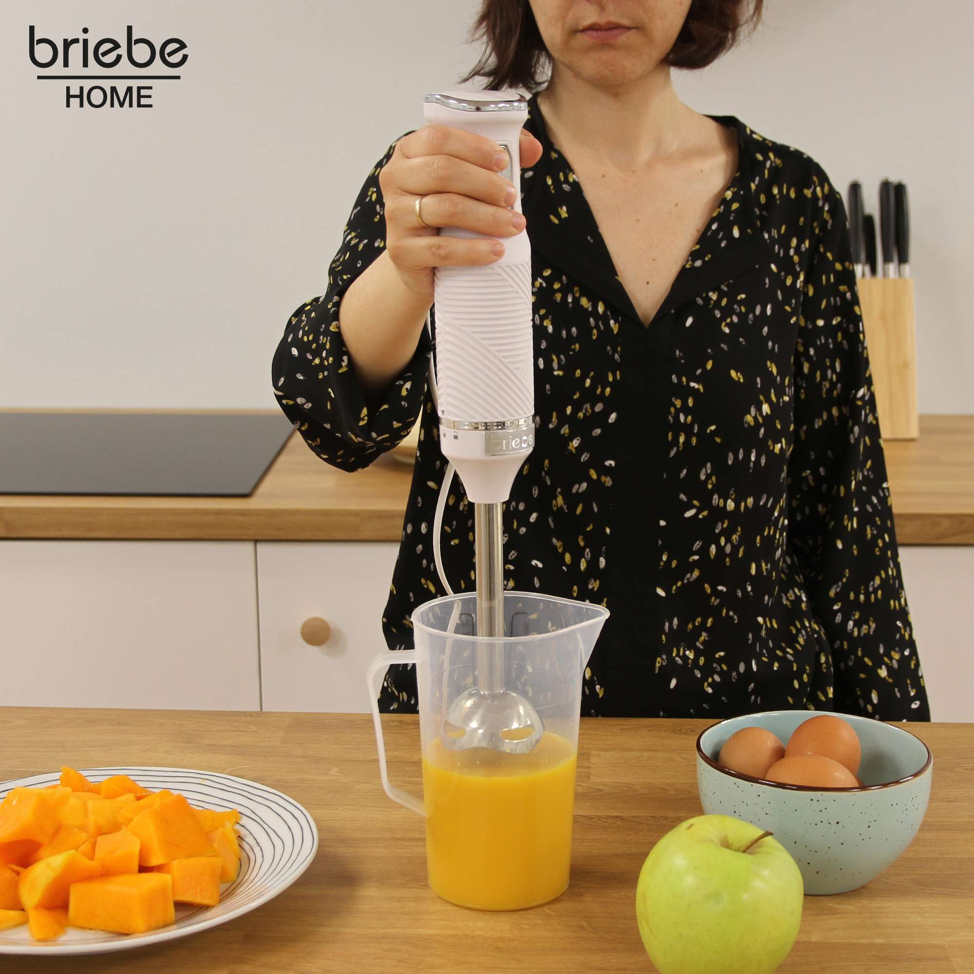 Una mujer usando una batidora de inmersión Briebe Home blanca para mezclar líquido naranja en una jarra. Manzana verde y huevos cerca.