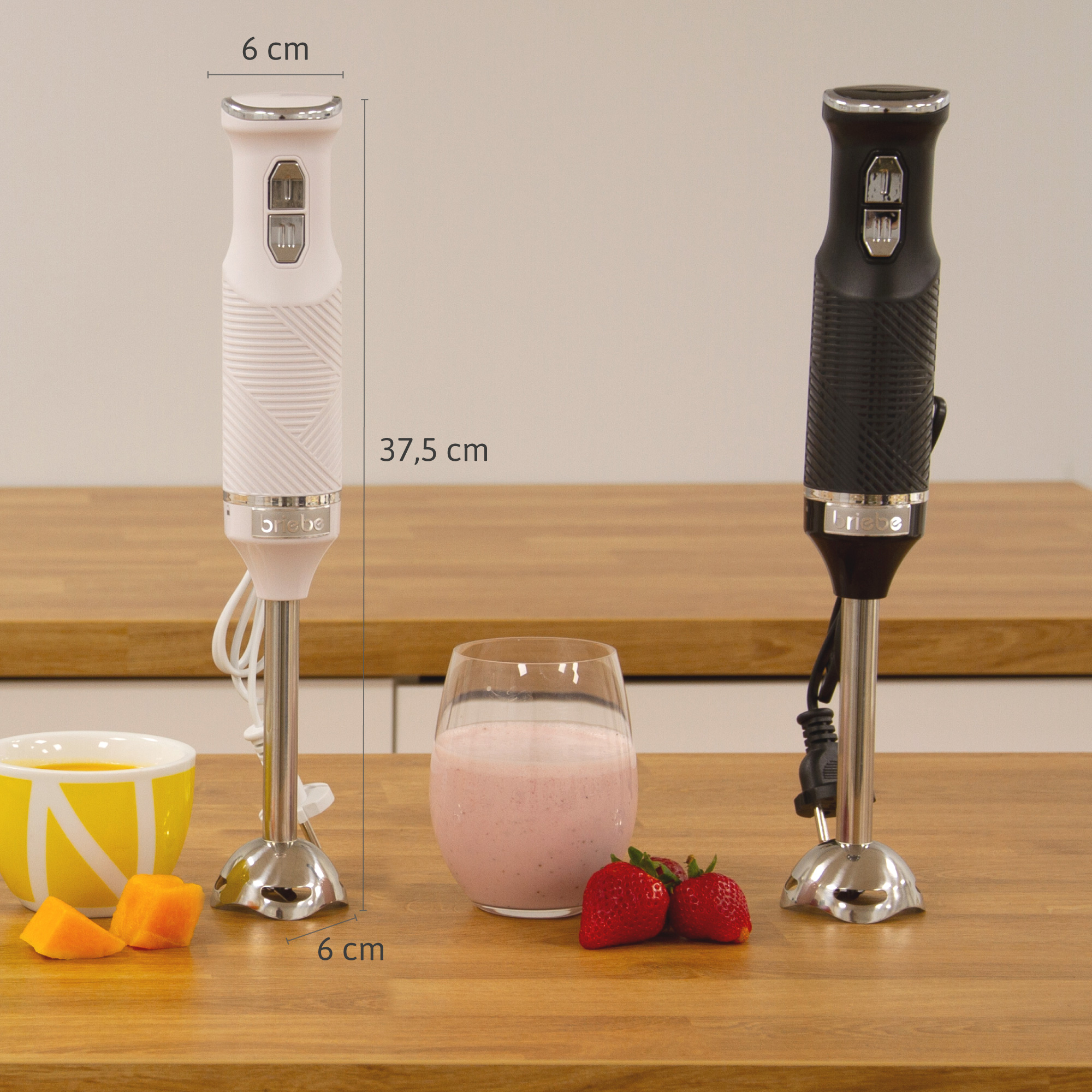 Dos batidoras de mano con un vaso de bebida rosa, fresas y un cuenco con fruta sobre una mesa de madera.