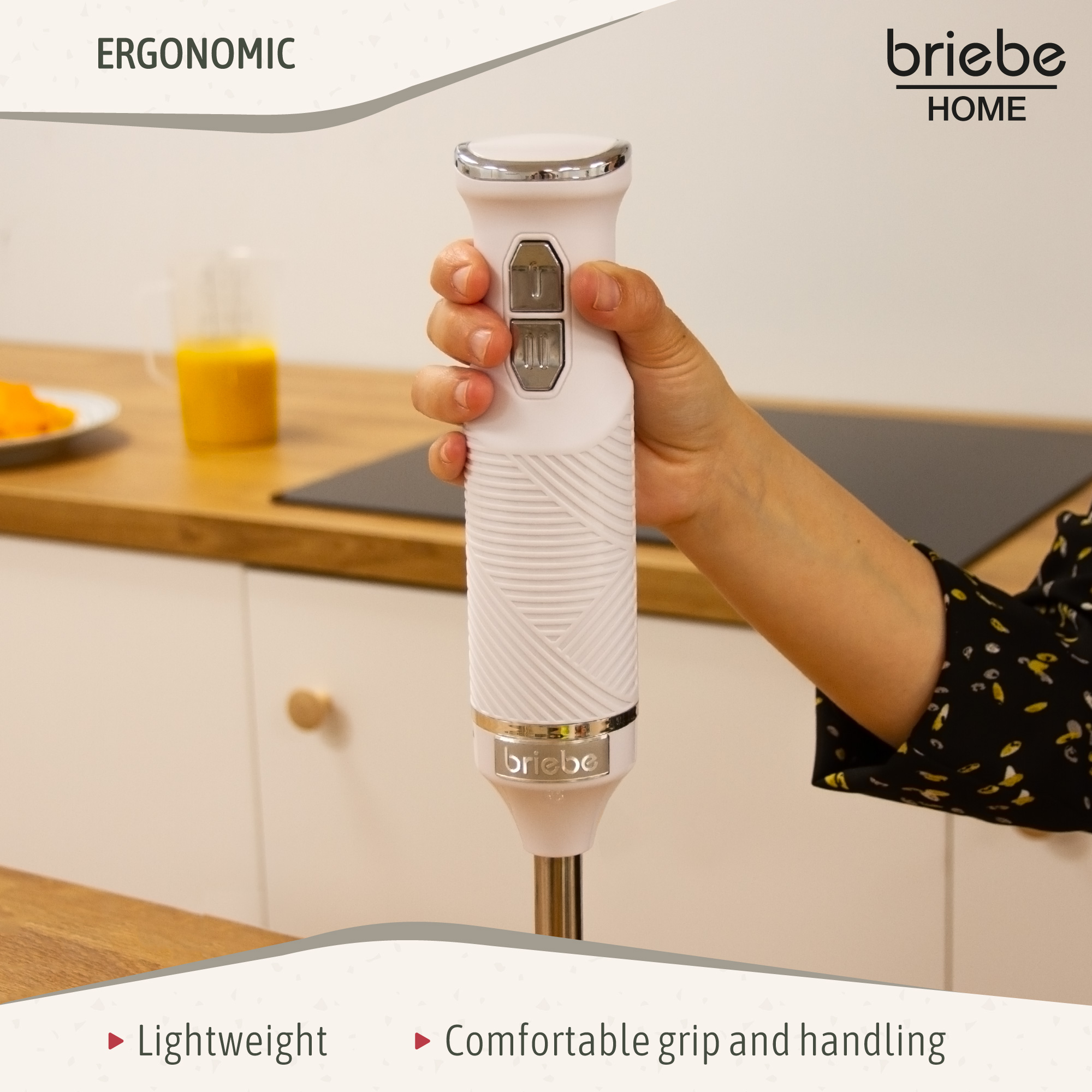 Una mano sosteniendo una batidora de inmersión Briebe Home blanca. El fondo muestra una encimera con un vaso de zumo.