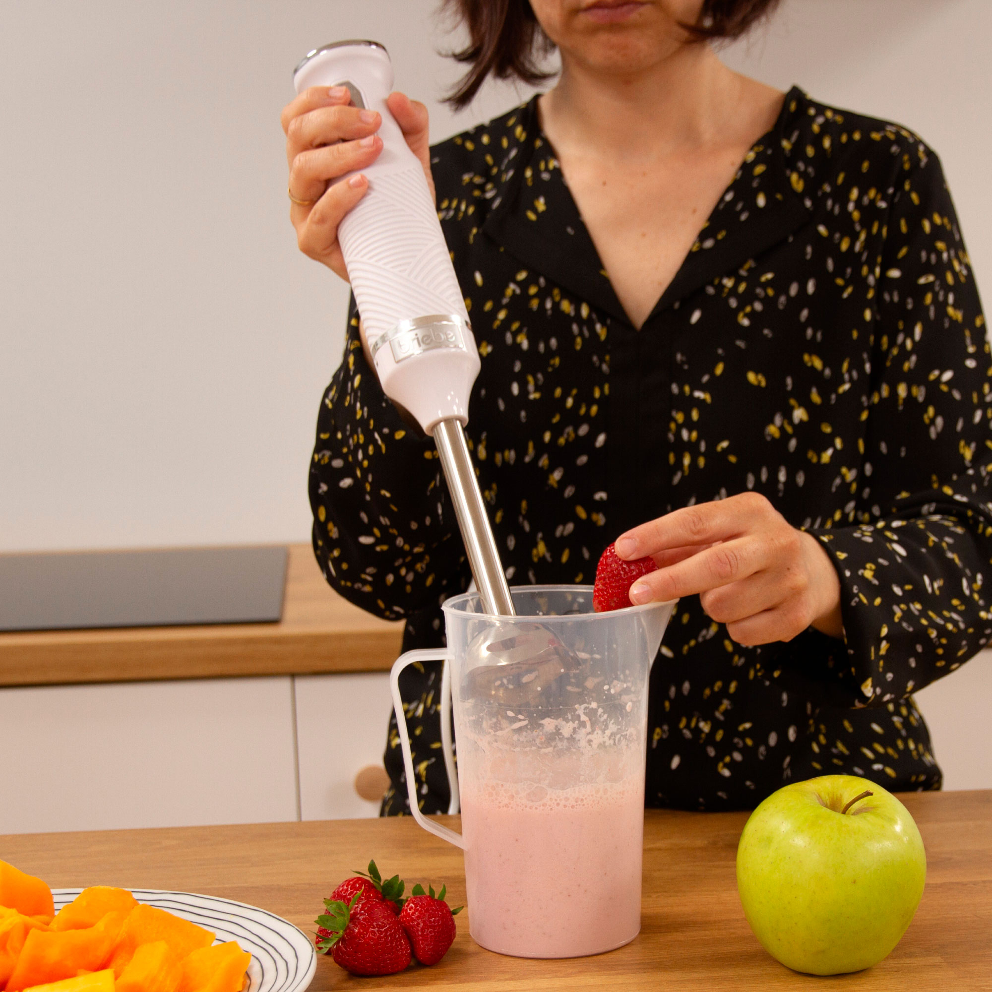 Una mujer usando una batidora de mano para hacer un batido rosa. Una manzana verde y fresas en la mesa.