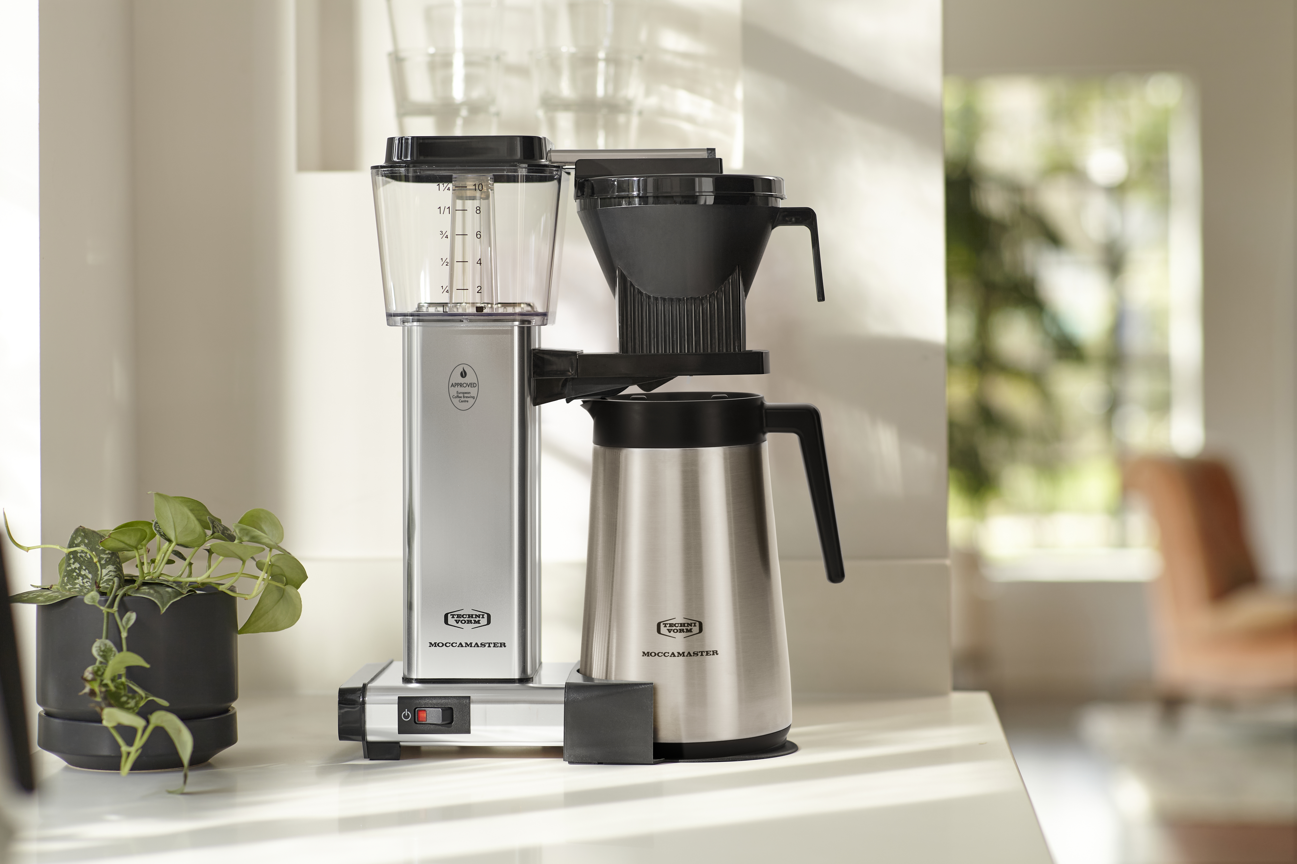 Une cafetière, corps argenté, dessus noir et carafe en verre. C'est sur un comptoir blanc.
