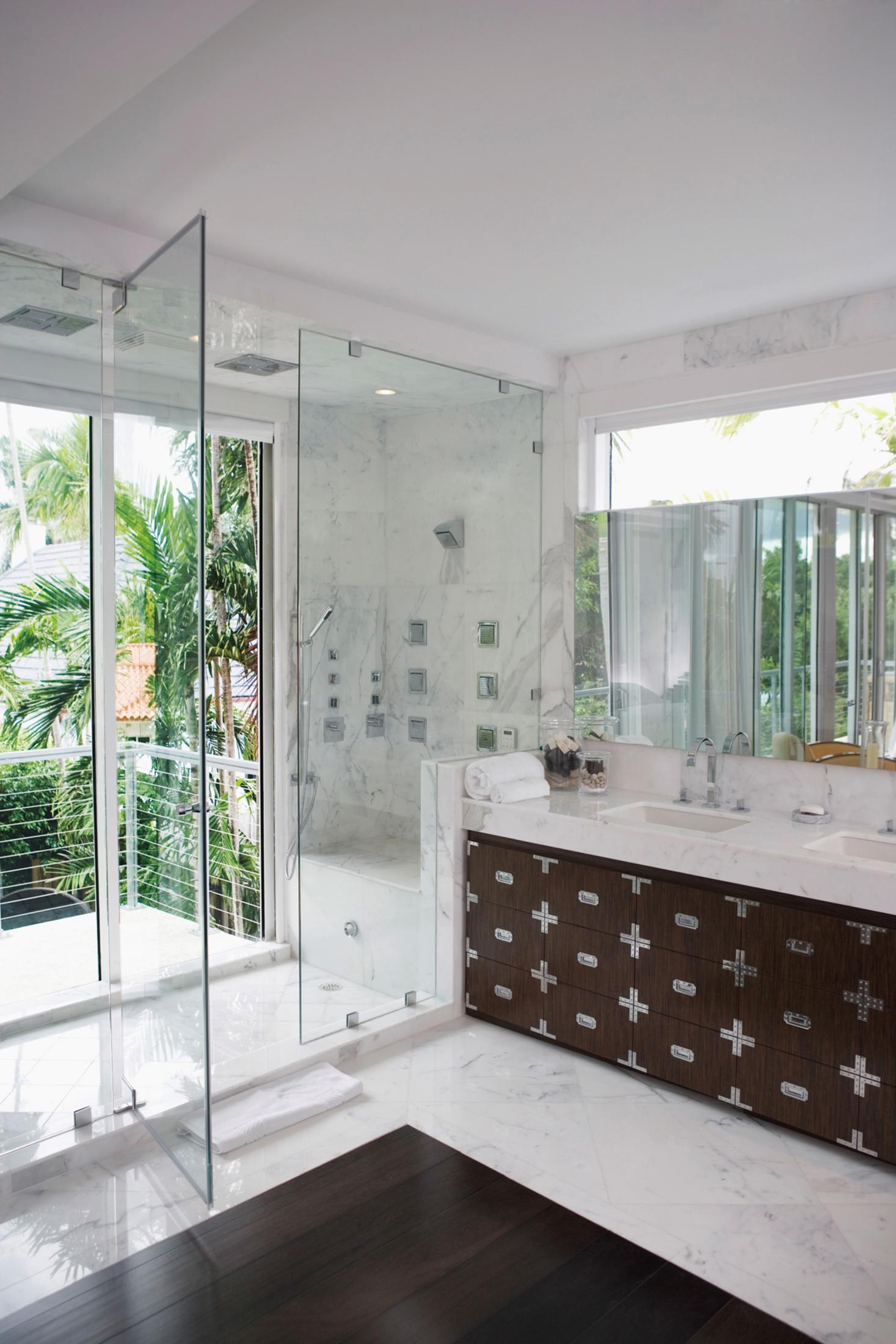 Cam duşakabinli, mermer duvarlı ve gümüş donanımlı ahşap dolaplı modern banyo.