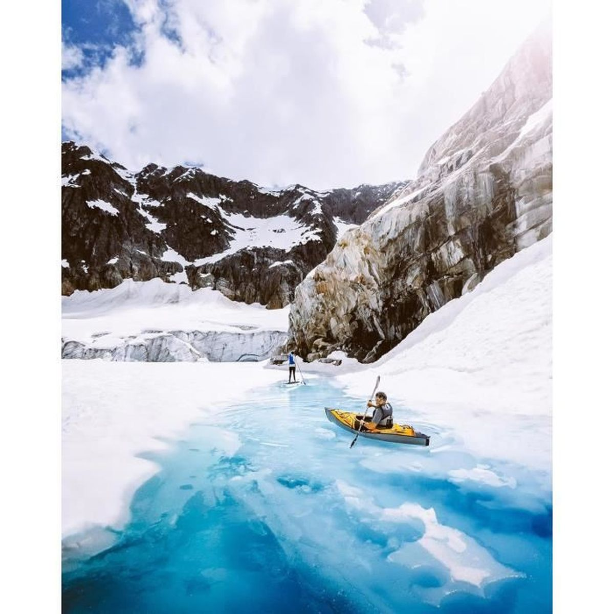 Eine Person fährt Kajak in einem blauen See, im Hintergrund schneebedeckte Berge. Der Himmel ist bewölkt.