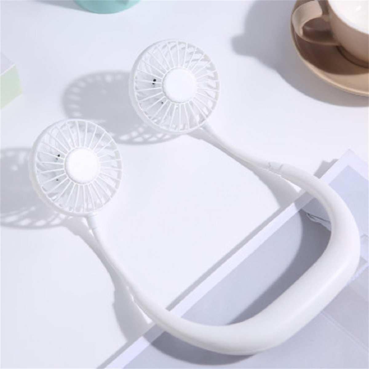 Zwei weiße persönliche Ventilatoren, verbunden mit einem weißen Nackenband. Sie befinden sich auf einer weißen Oberfläche.