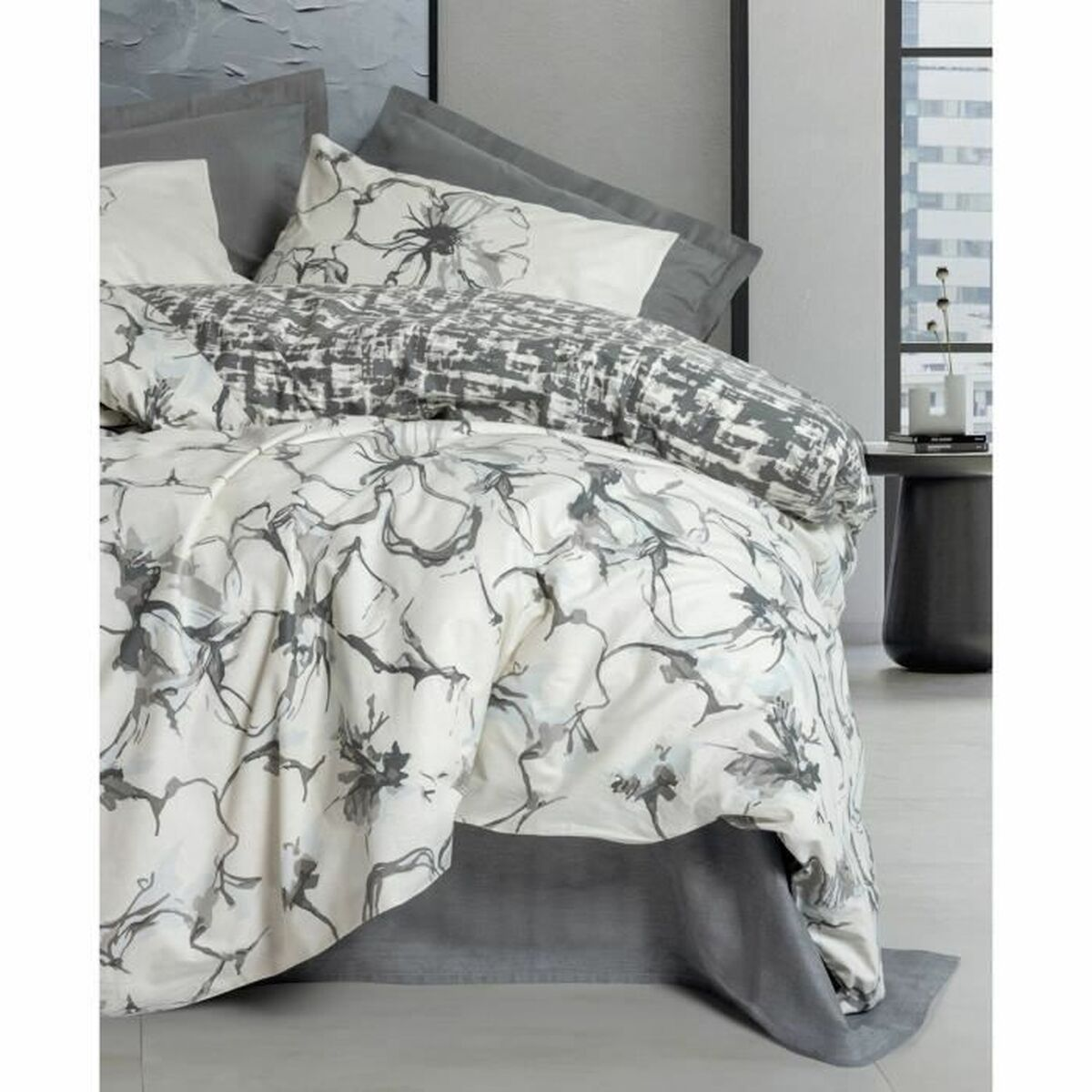 Weißes Bettwäsche-Set mit grauem Blumendesign. Kissen und Bettdecke sichtbar. Hintergrund ist ein Raum mit Fenstern.