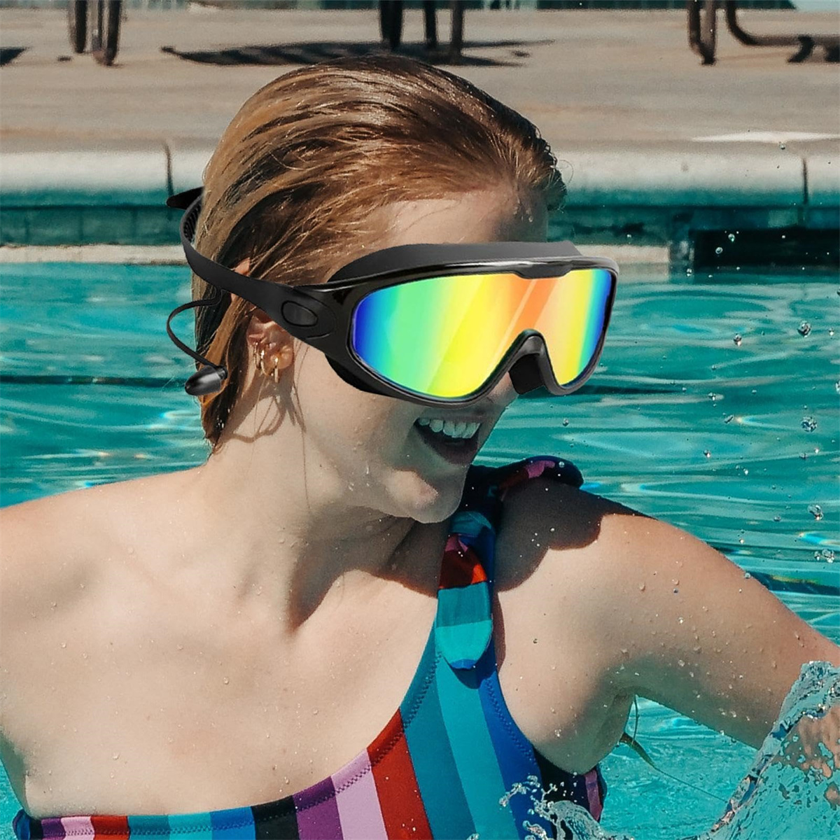 Frau im Pool, die eine Schwimmbrille mit Regenbogenlinsen trägt und lächelt, während Wasser um sie herum spritzt.