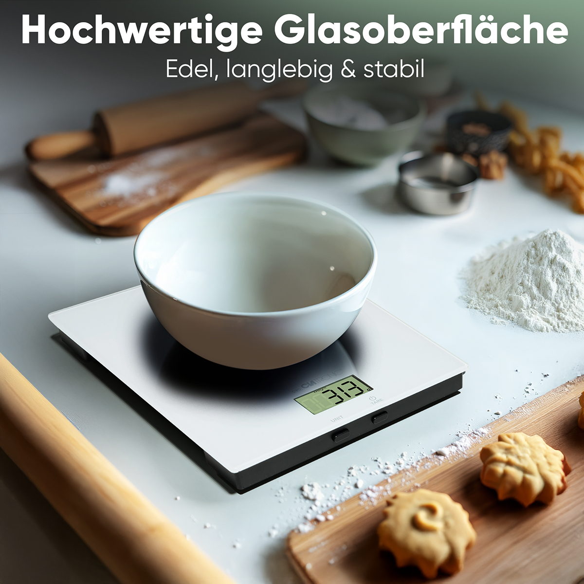 Eine weiße Waage mit einer Schüssel darauf. Mehl und Küchenutensilien liegen auf der Oberfläche.