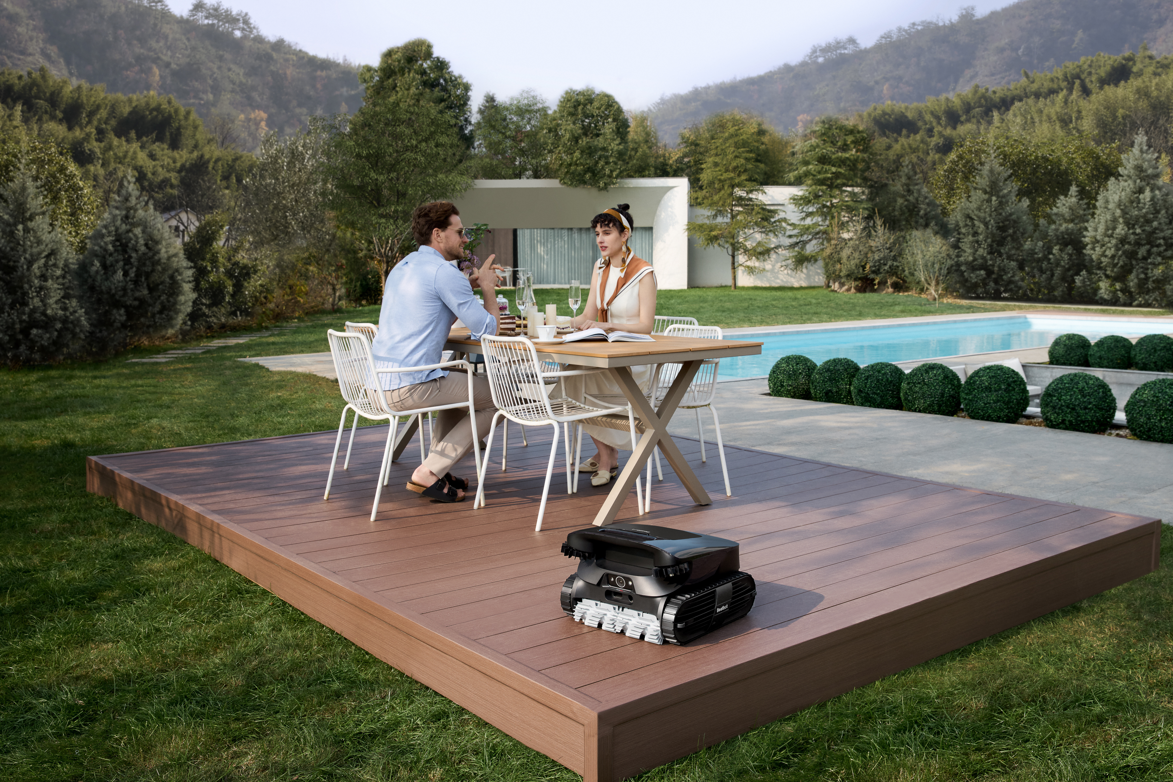 Una pareja sentada en una mesa en una terraza, un cortacésped robótico en la terraza. Árboles y una piscina en el fondo.