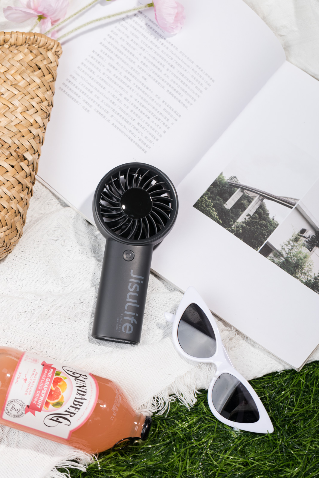 Ein schwarzer Jisulife-Ventilator, Sonnenbrille, eine Flasche Saft, eine Strohtasche und ein offenes Buch auf weißem Stoff.