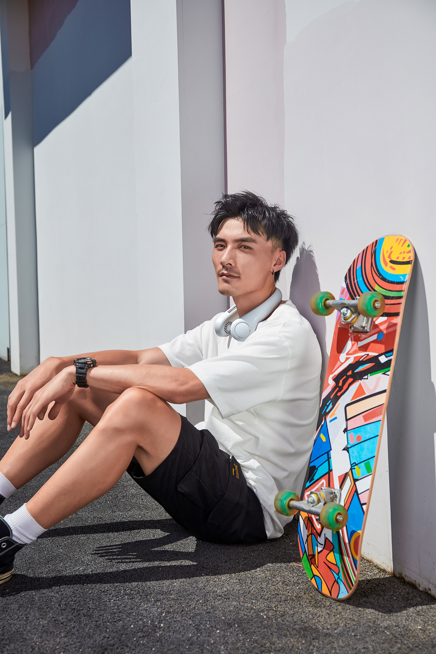 Ein Mann sitzt an einer Wand mit einem Skateboard. Er trägt ein weißes Hemd und schwarze Shorts.