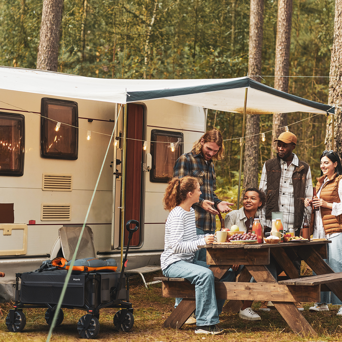 A group of people at a campsite with a wagon. They're around a picnic table near a camper.