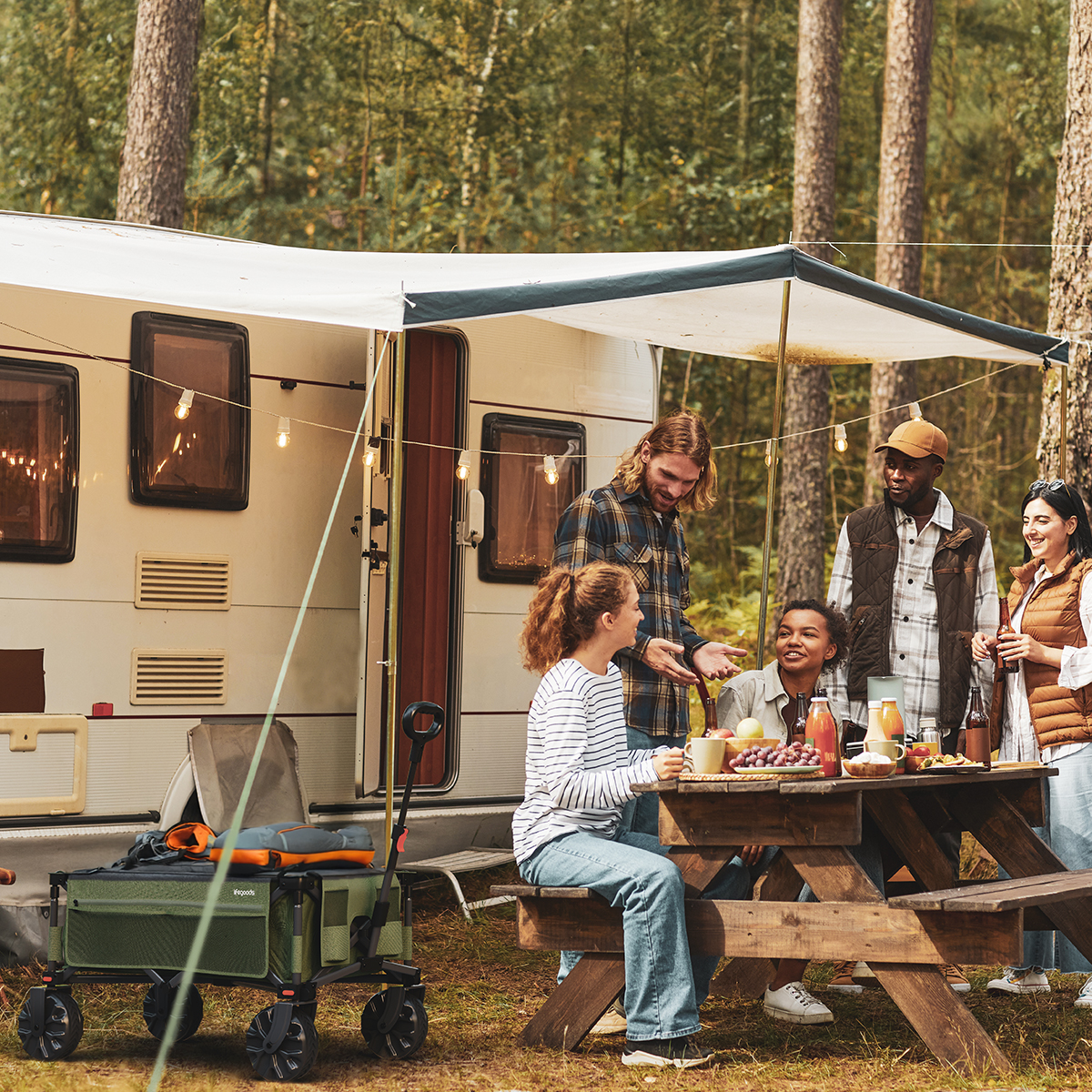 People at a picnic table near a caravan. A wagon with green fabric and large wheels is in the foreground.