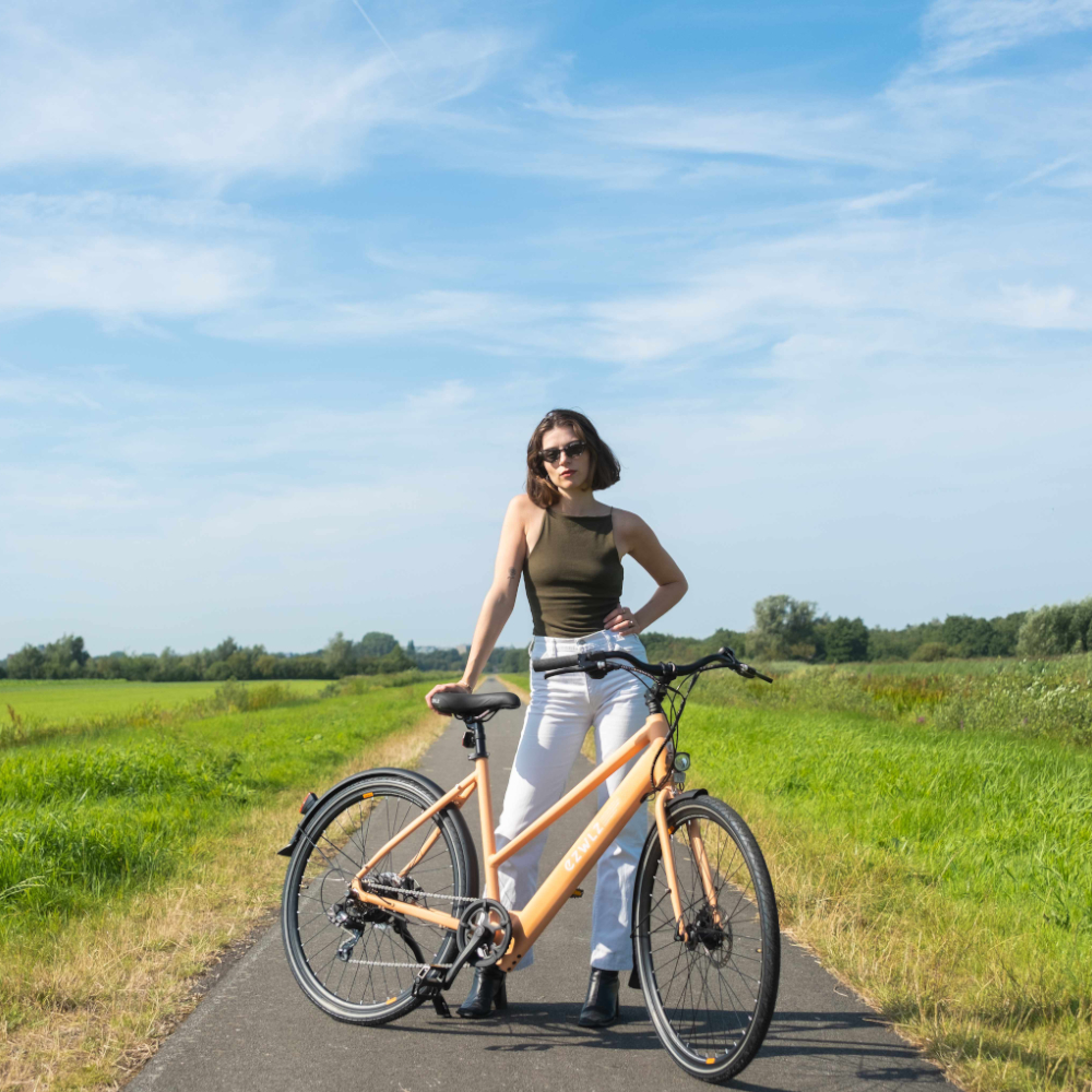 Vrouw staat met oranje e-bike op een weg, met groene velden en blauwe lucht op de achtergrond.