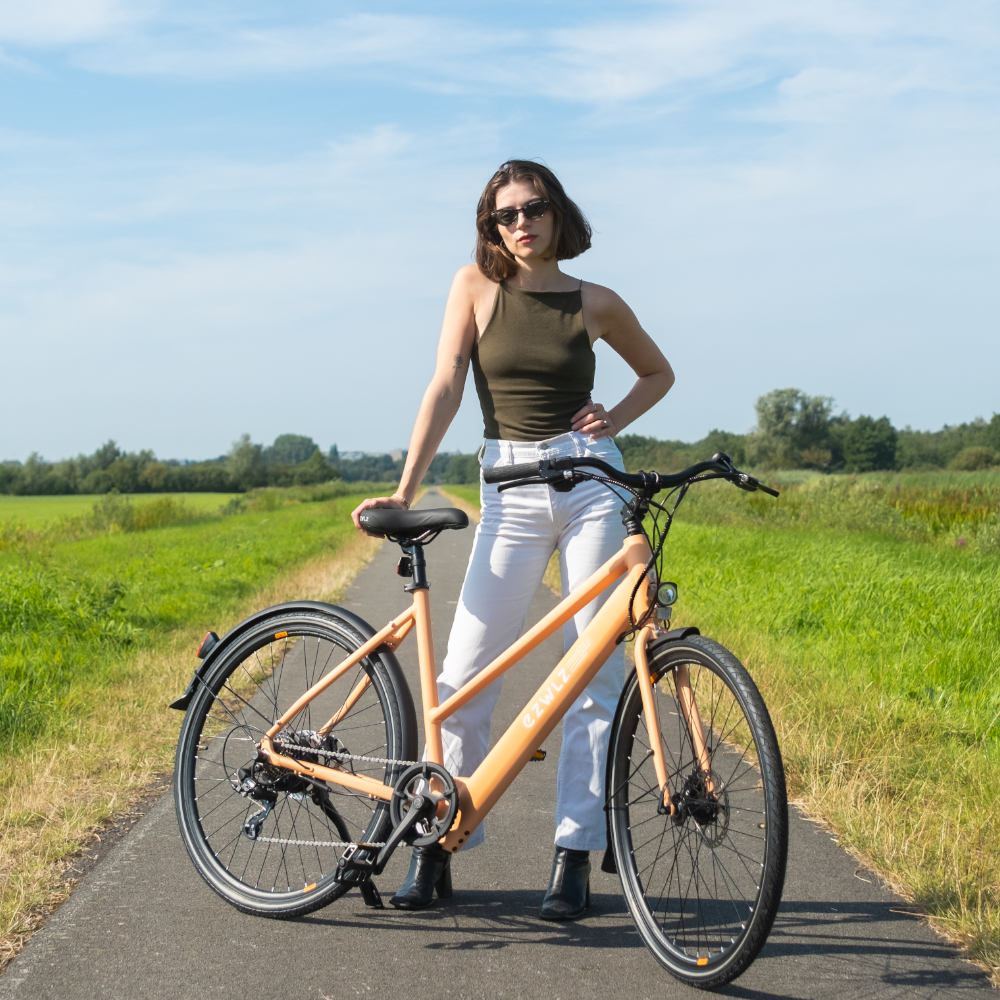 Vrouw met zonnebril staat met oranje fiets op pad, velden op achtergrond.