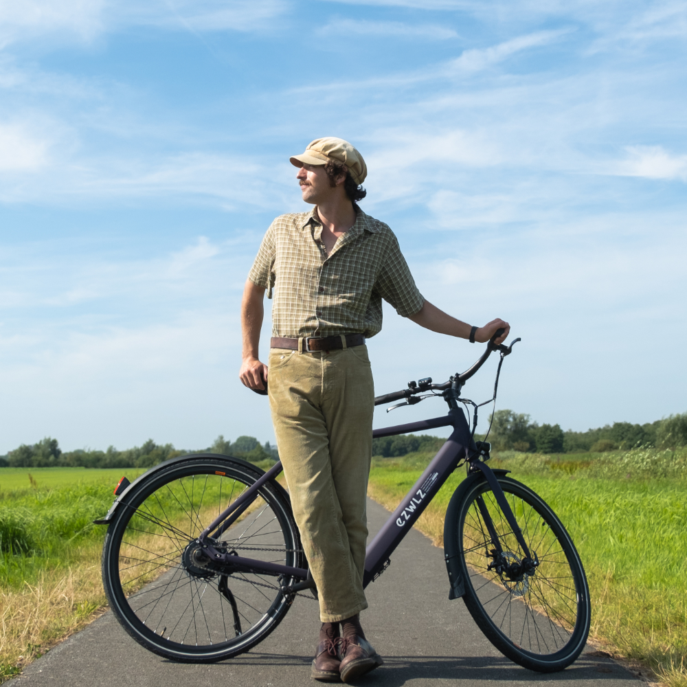 Man met pet staat met paarse fiets op pad, veld op achtergrond.