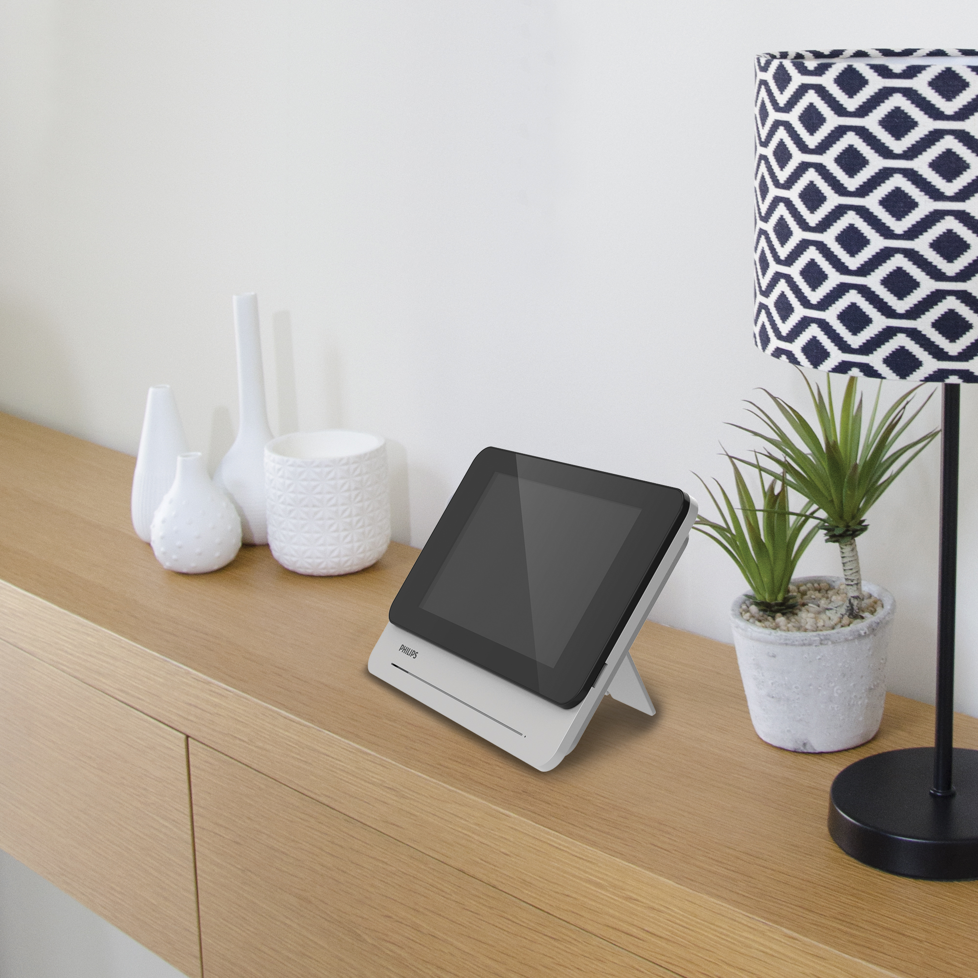 Un dispositivo tipo tableta se muestra en una superficie de madera. Tiene el logotipo de 'Philips'. Hay elementos decorativos y una lámpara.