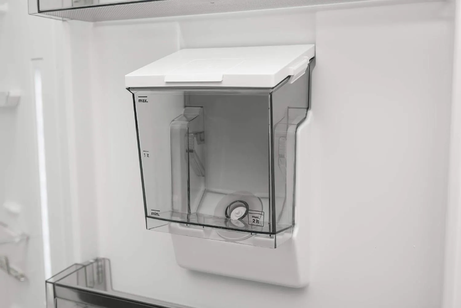 Primo piano di un serbatoio d'acqua in plastica trasparente all'interno di un frigorifero bianco, che mostra gli indicatori del livello di riempimento.