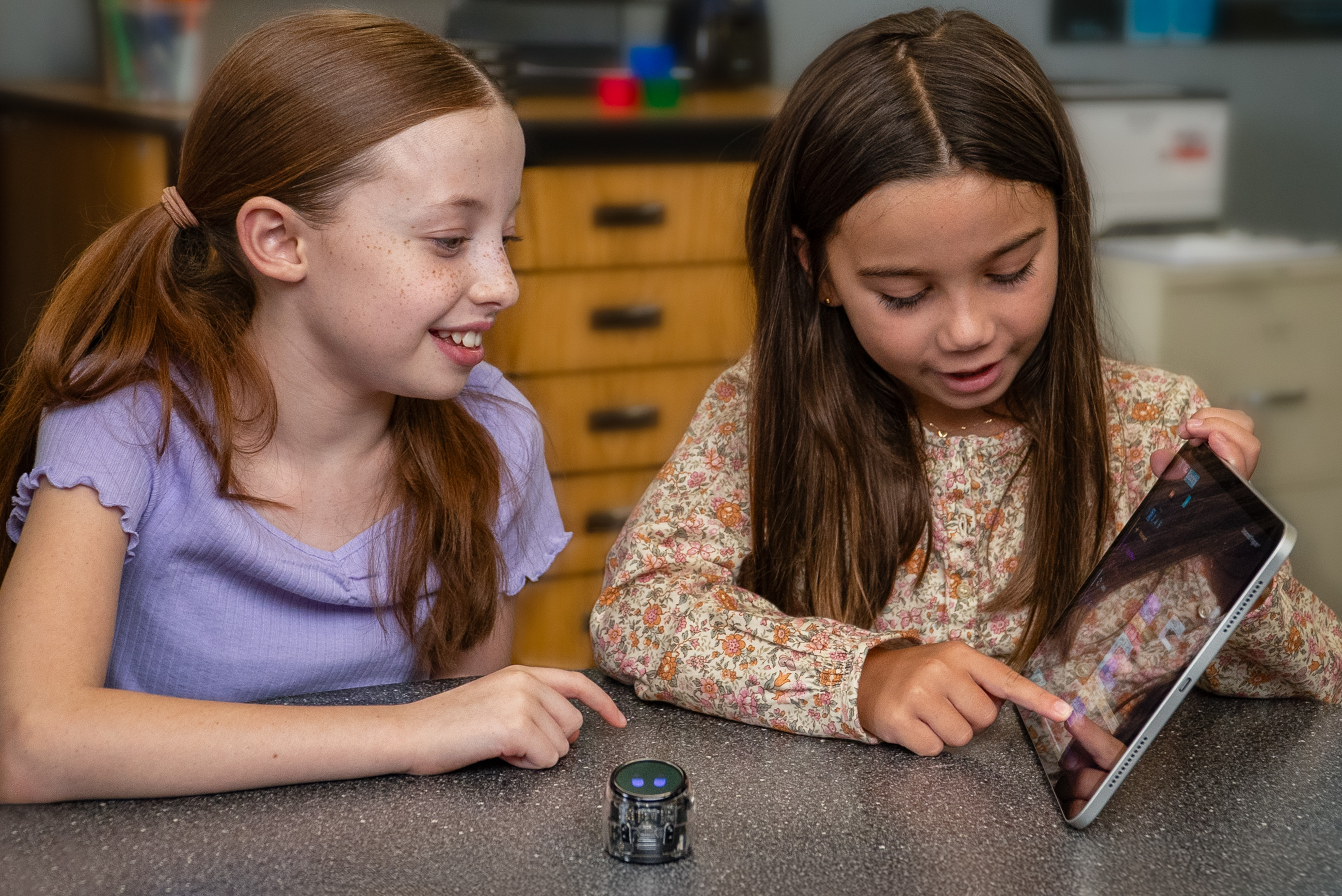 Two young girls look at a tablet. A small robot sits on the table. The girls are smiling.