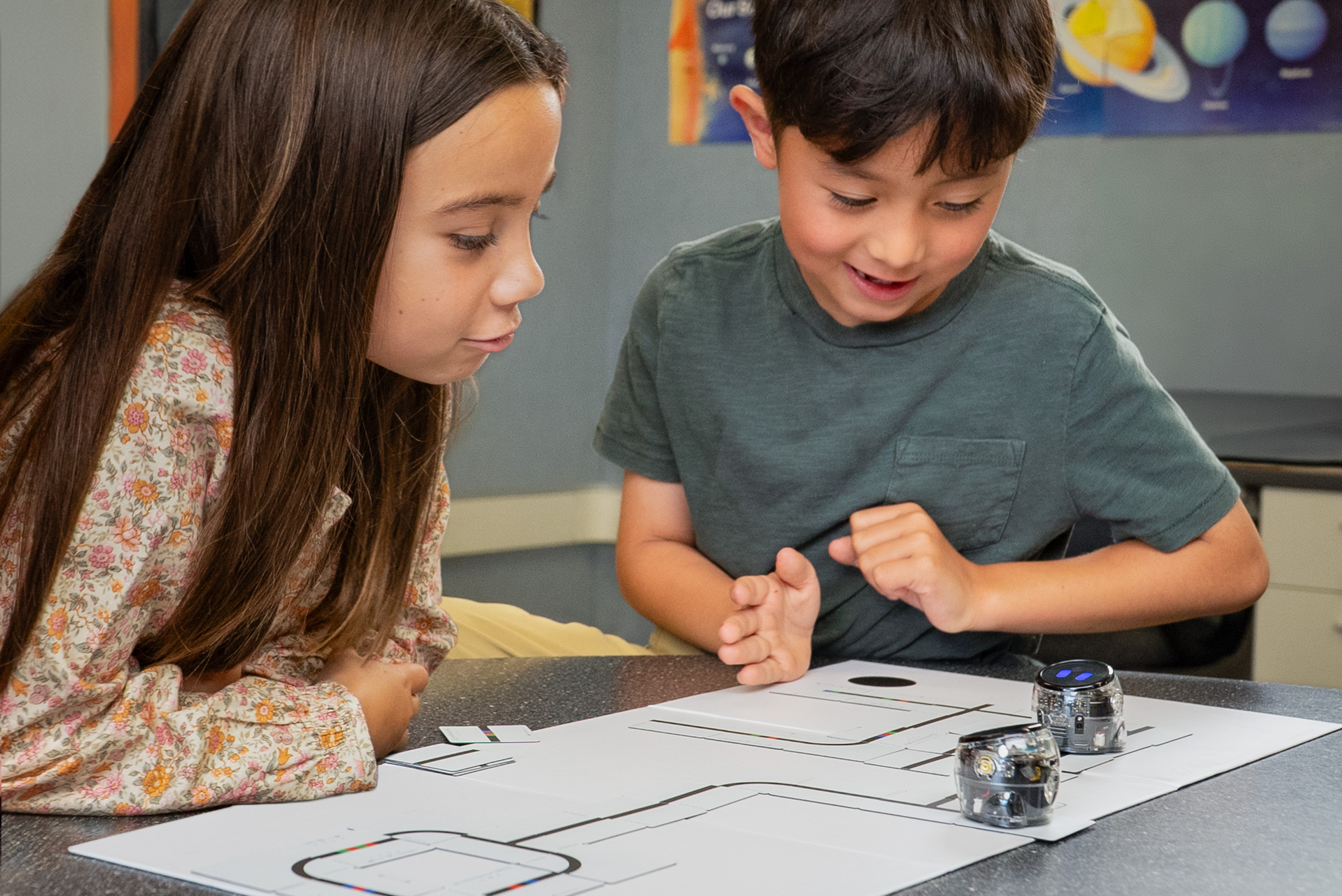 Two robots follow a line on a printed sheet. A boy and girl watch them closely and smile.