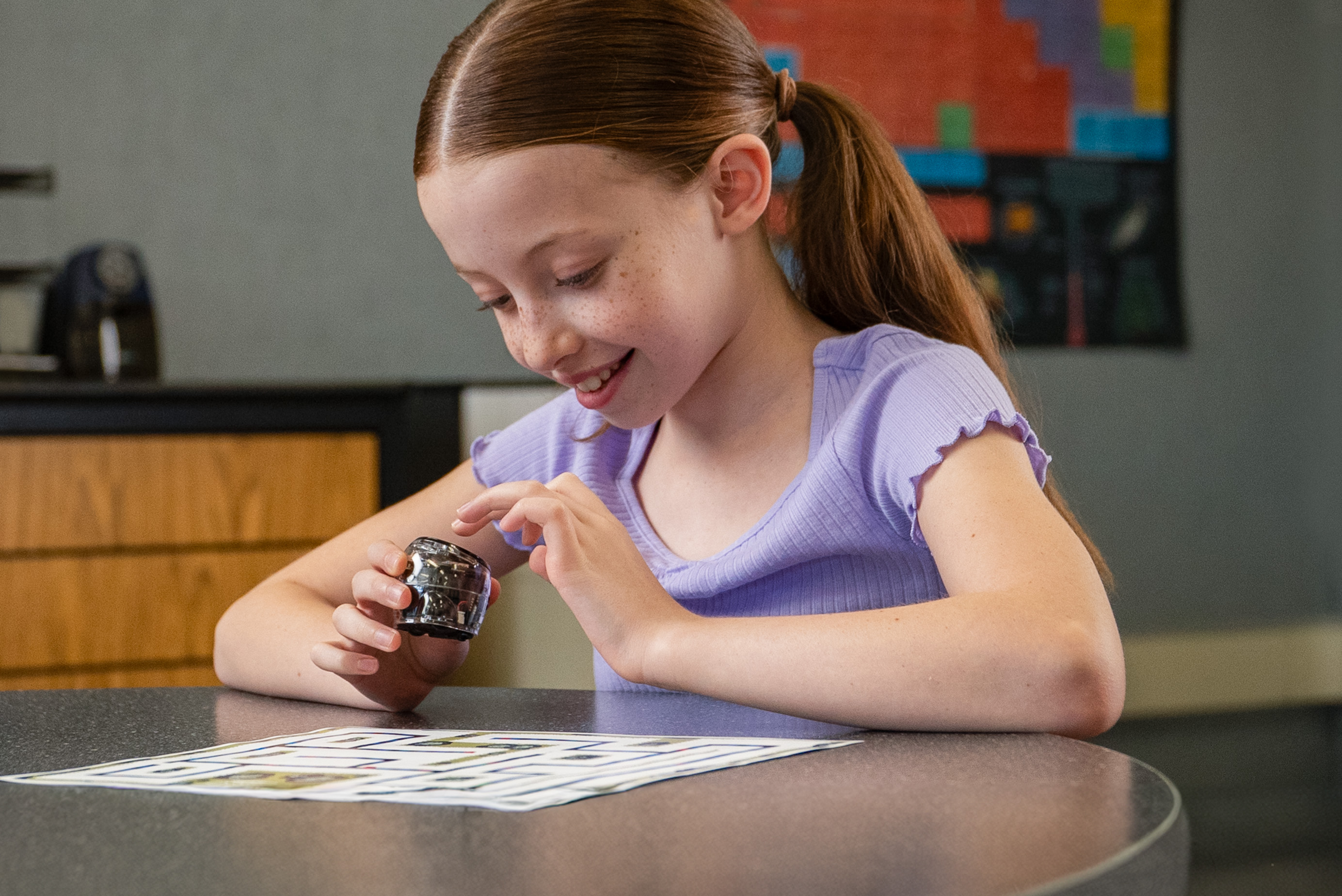 A girl smiles at a small robot on a table. A printed sheet with lines is in the foreground.