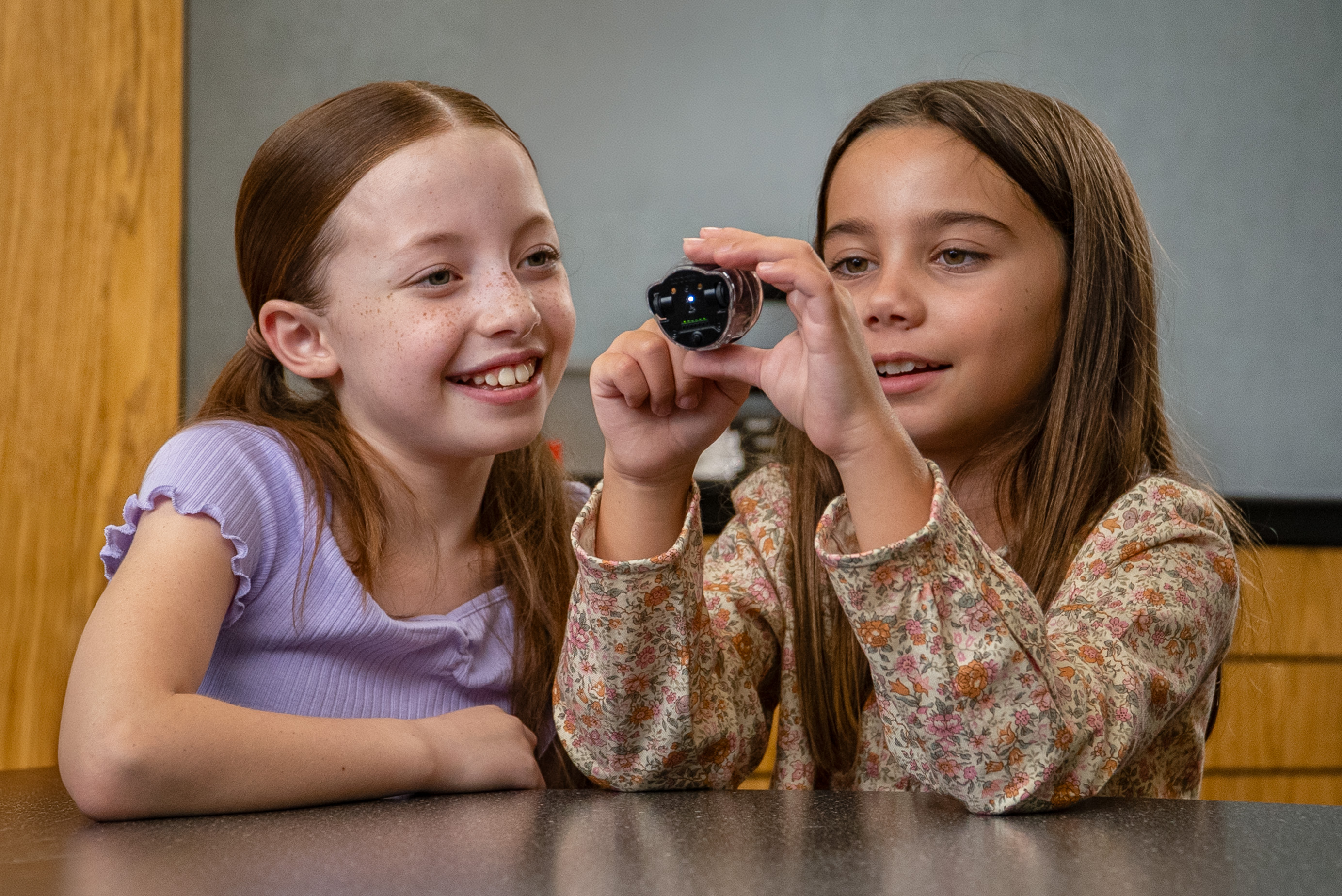 Two girls are looking at a small robot, smiling.