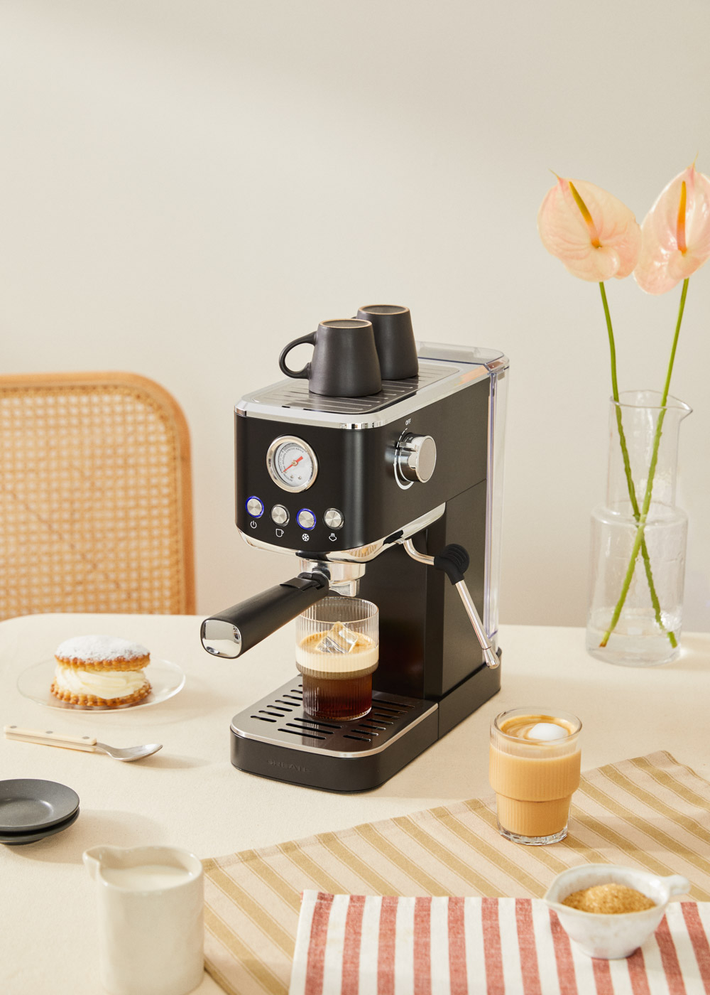 Máquina de espresso negra con café y tazas en una mesa con flores y comida.