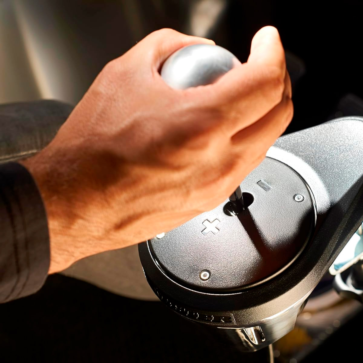 Une main agrippe un pommeau de levier de vitesse métallique. Le levier de vitesse comporte des symboles plus et moins et est entouré de matériau noir.