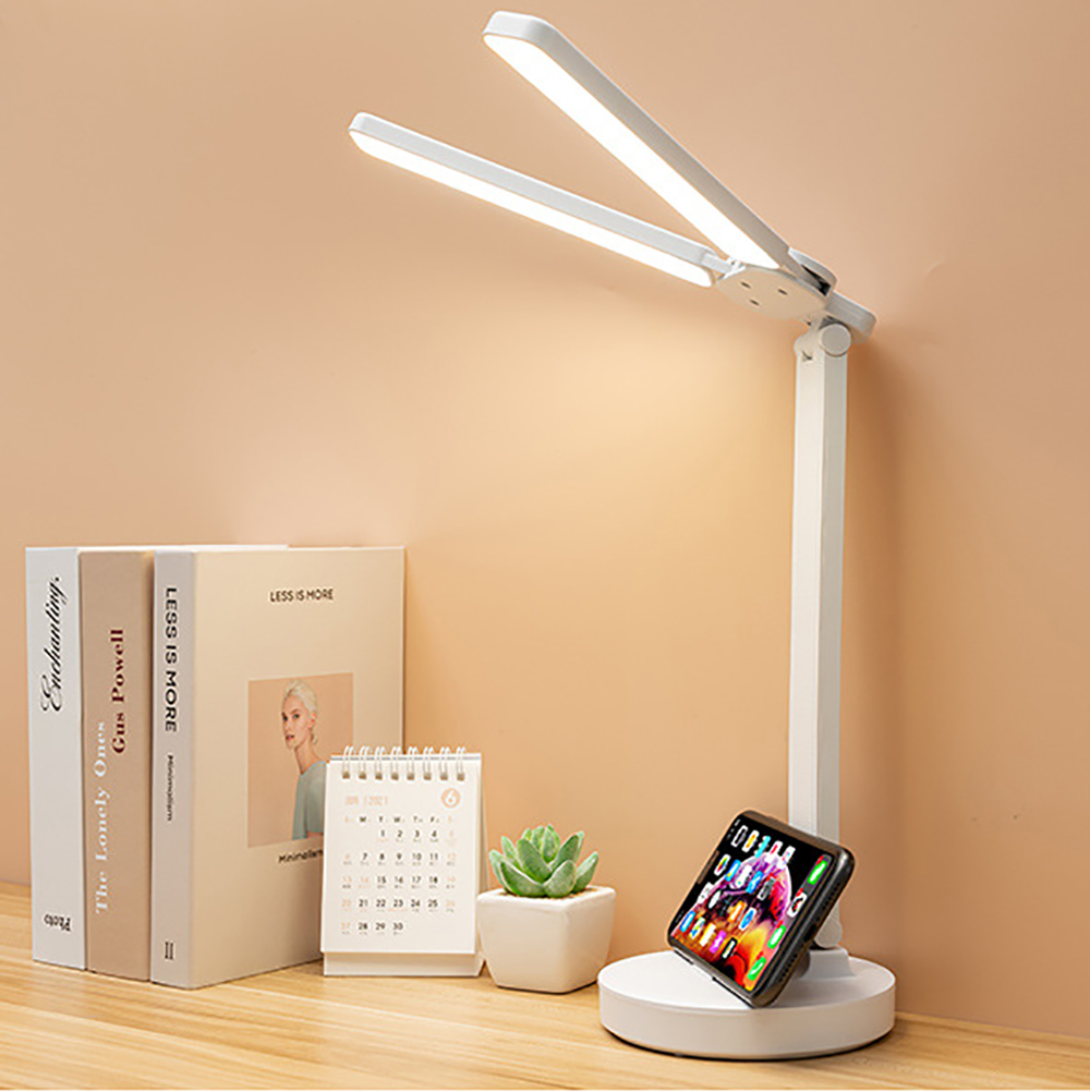 Eine weiße Schreibtischlampe mit verstellbaren Lichtpaneelen und ein Telefon, das lädt.