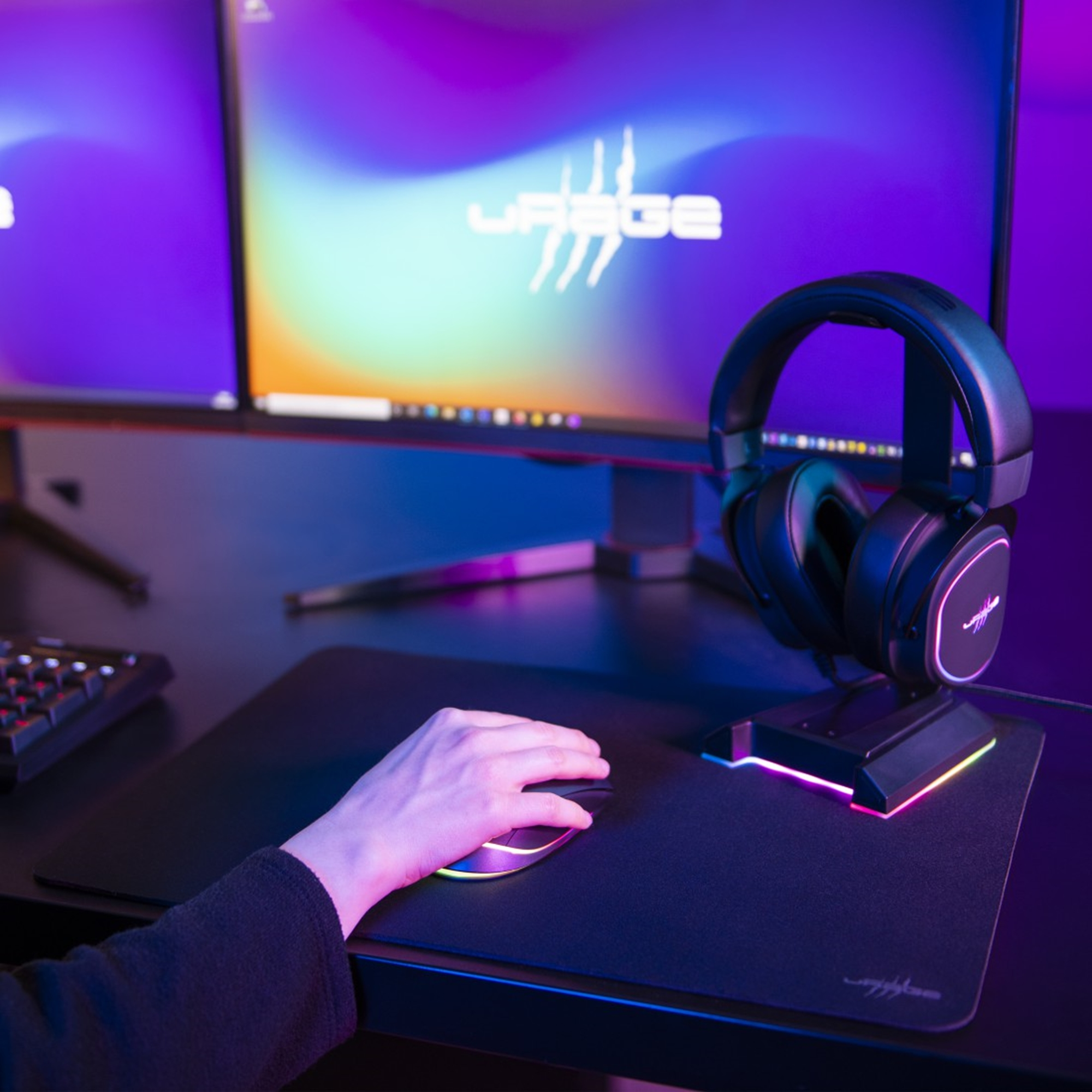 Eine Hand mit einer Gaming-Maus auf einem Mauspad, im Hintergrund ein Monitor und ein Headset.