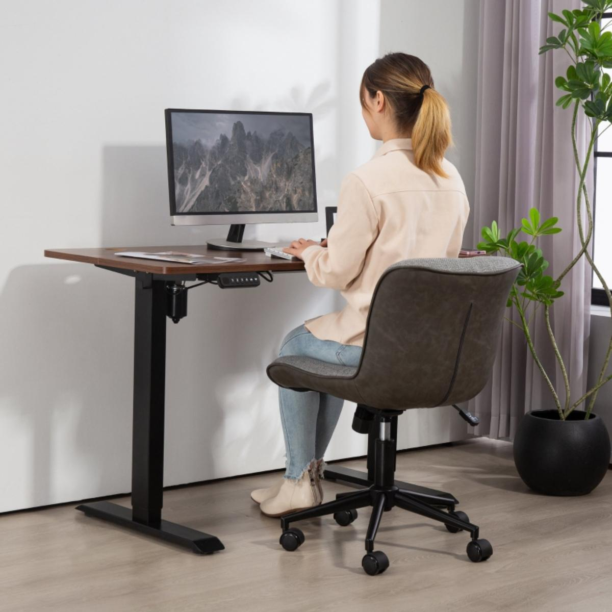 Eine Frau sitzt an einem höhenverstellbaren Schreibtisch und arbeitet am Computer. Stuhl und Pflanze in der Nähe.