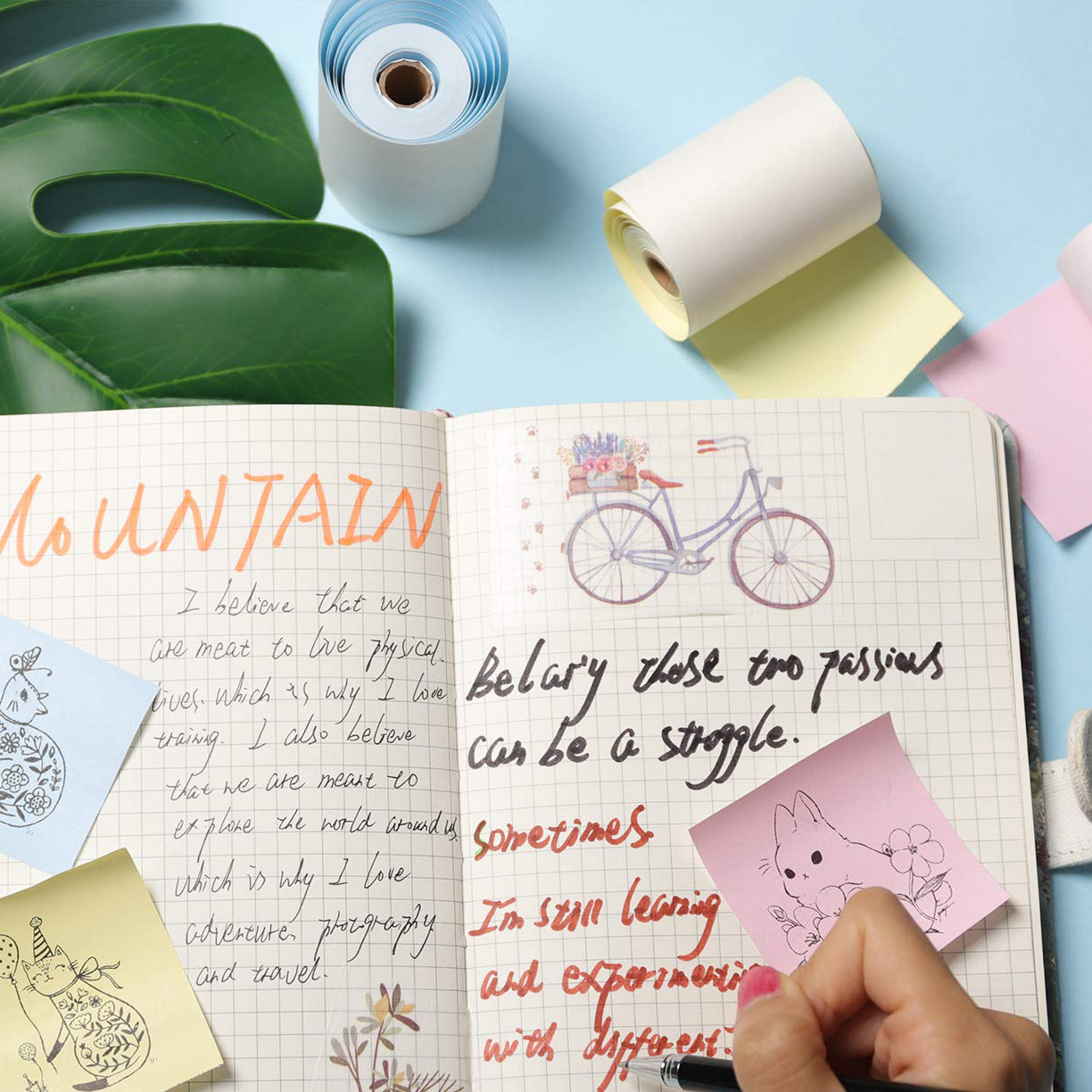 Ein Notizbuch mit handschriftlichen Notizen und Aufklebern, sowie eine Fahrradzeichnung. Papierrollen und Stifte liegen daneben.