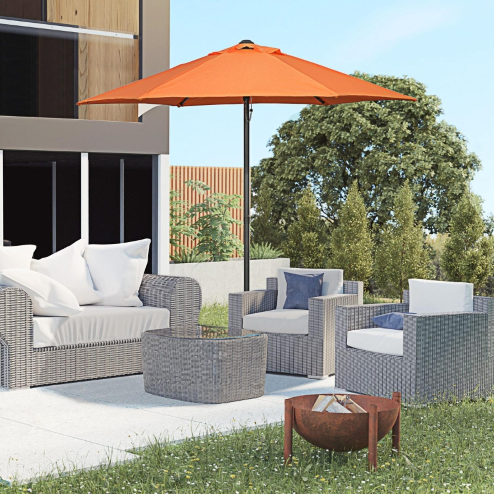 Szene im Freien: Terrasse mit Korbmöbeln, orangefarbenem Sonnenschirm und Feuerstelle. Grünes Gras und Bäume im Hintergrund.