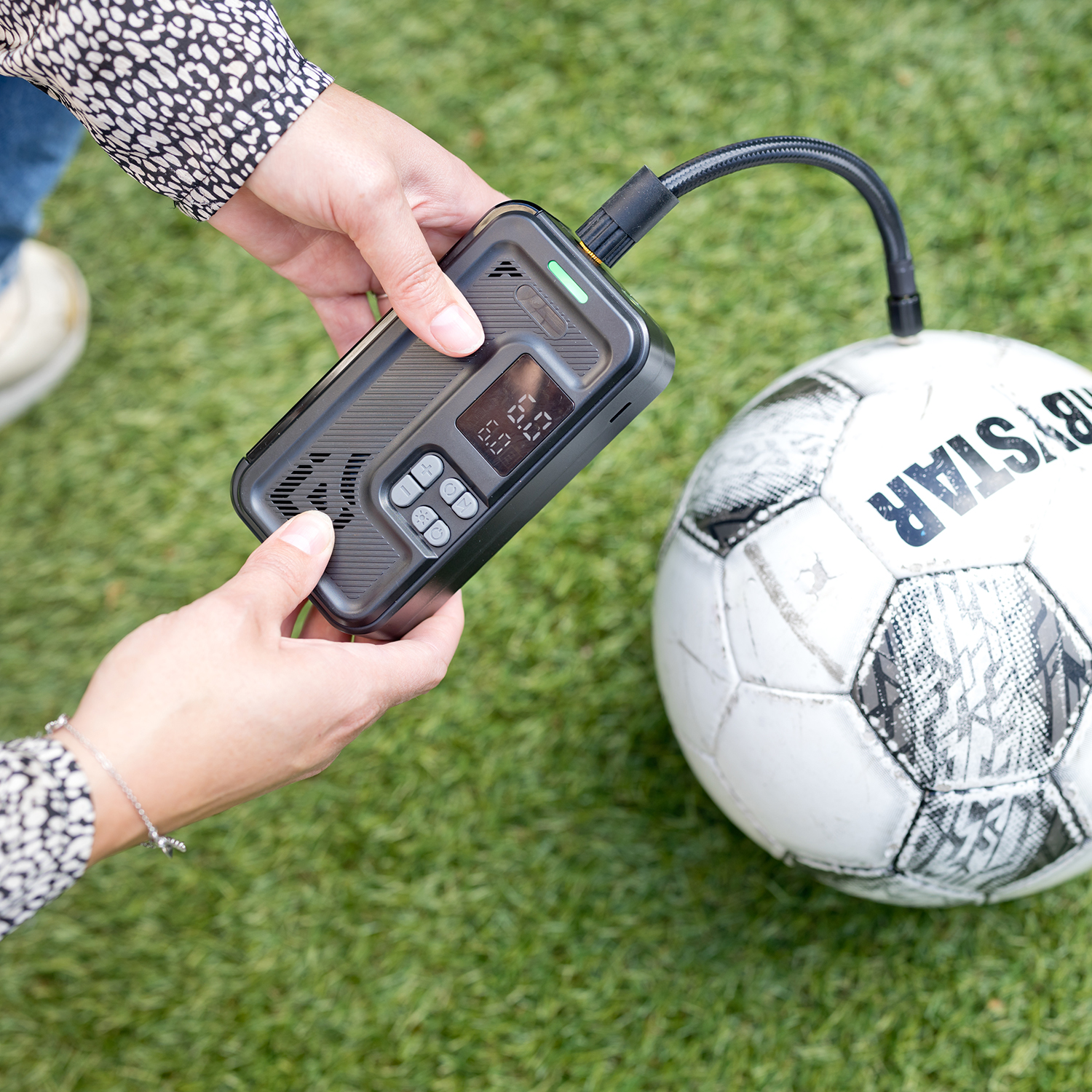 Handen pompen een voetbal op met een zwart apparaat op groen gras.
