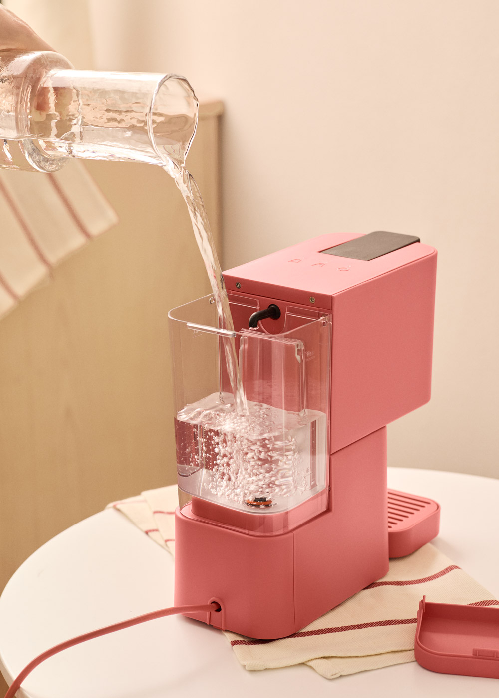 Una persona vierte agua de una jarra de vidrio en una cafetera rosa. La máquina está sobre una mesa blanca.