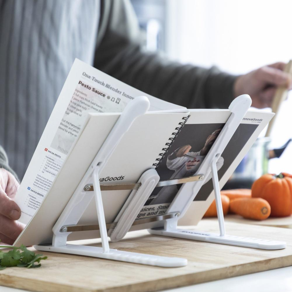 Un leggio per libri di cucina con un libro aperto, le mani di una persona sono visibili, vicino agli ingredienti e a un tagliere.