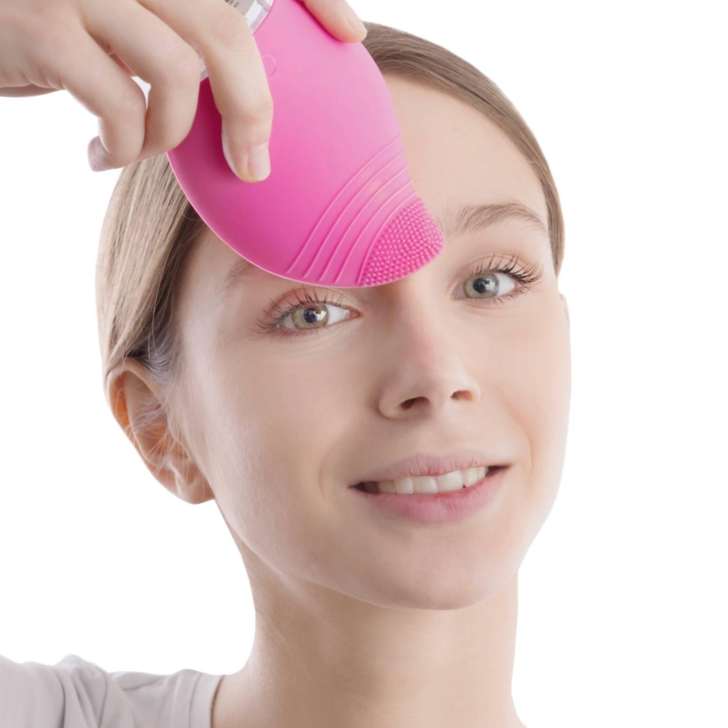 Donna che applica una spazzola rosa per la pulizia del viso sul suo viso, sorridendo.