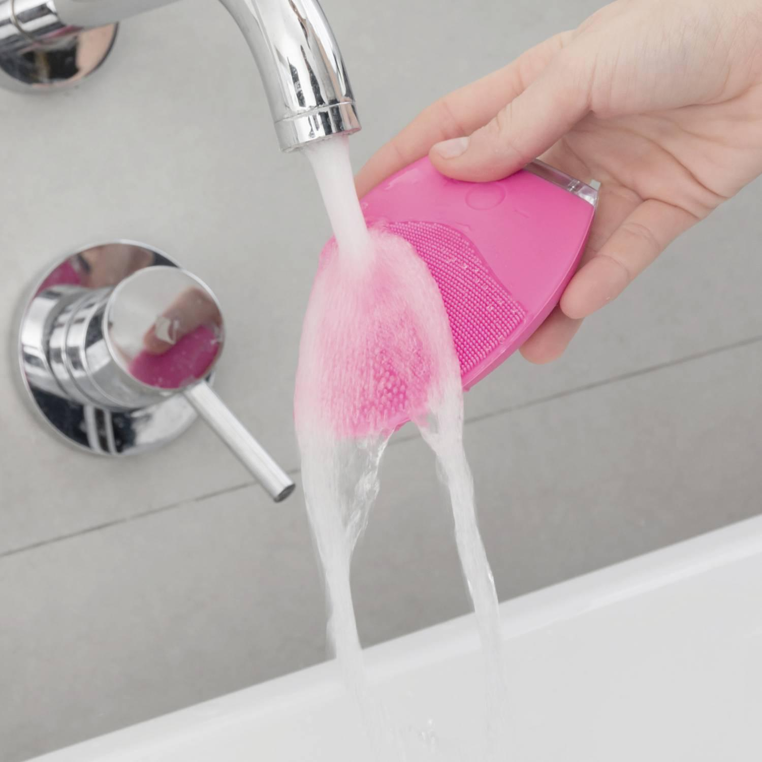 Una spazzola rosa per la pulizia del viso viene tenuta sotto l'acqua corrente di un rubinetto, pulendo la testina.