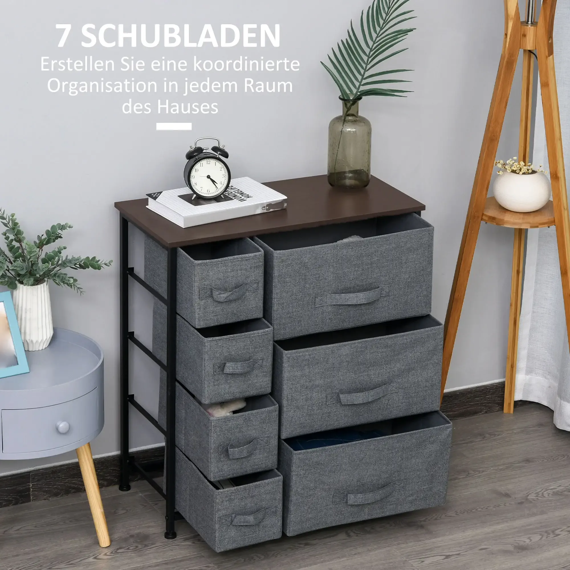 Eine Kommode aus grauem Stoff mit sieben Schubladen. Sie hat eine dunkle Holzplatte und ein Metallgestell. Eine Uhr steht darauf.