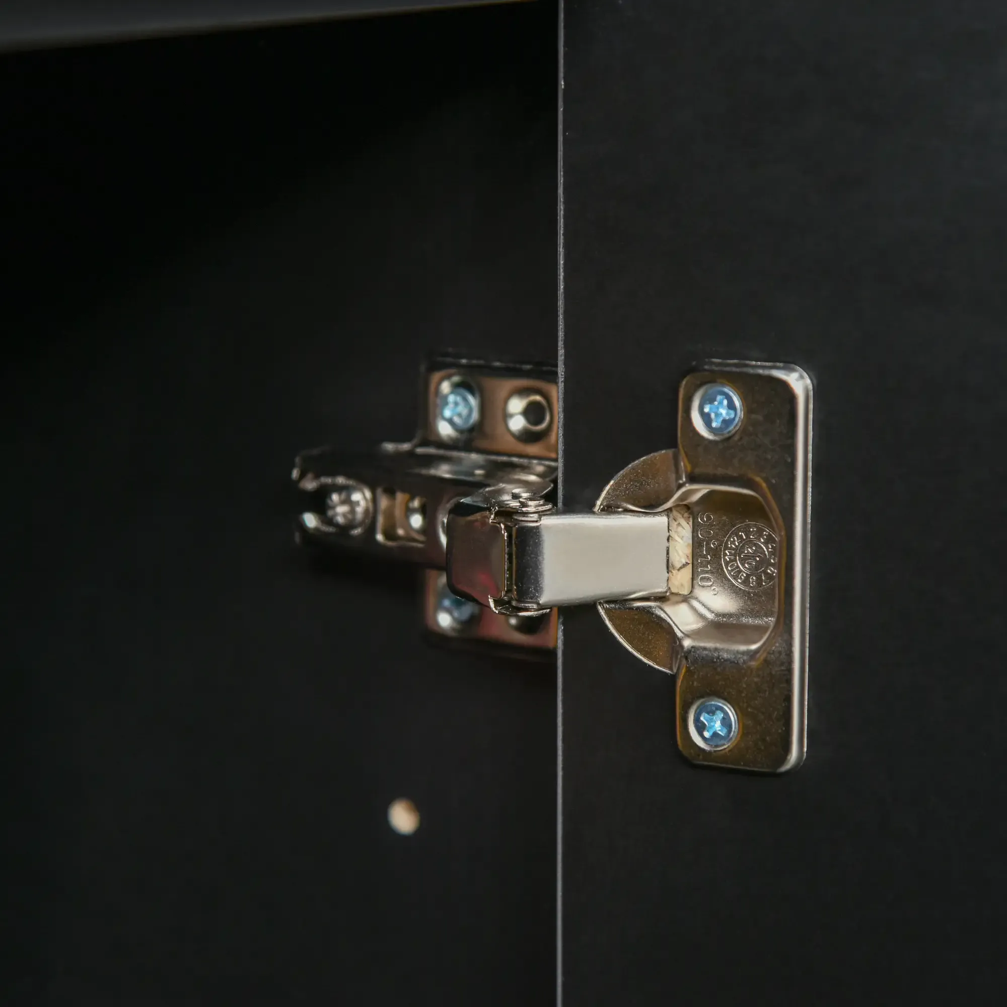 Nahaufnahme eines Schrank-Scharniers. Das Scharnier ist metallisch silbern und an einer schwarzen Schranktür befestigt.