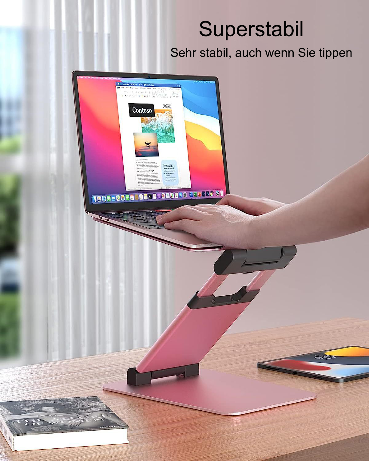 Laptop auf einem Ständer mit einem Buch und einem Tablet auf dem Schreibtisch. Rosa Ständer und Schreibtisch.
